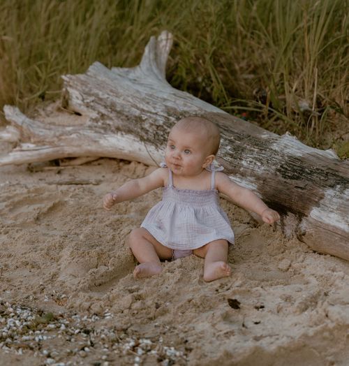 Baby in light gray dress sitting on sandy ground near driftwood log.