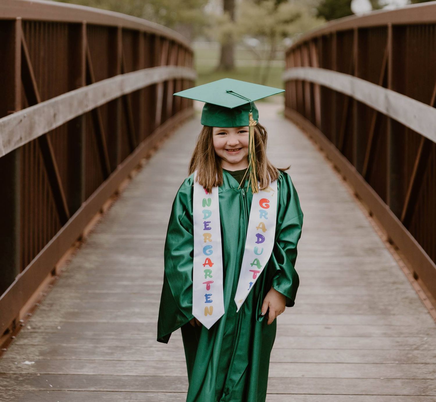 Graduate in emerald green cap and gown walking along wooden bridge pathway through trees for graduation photos.