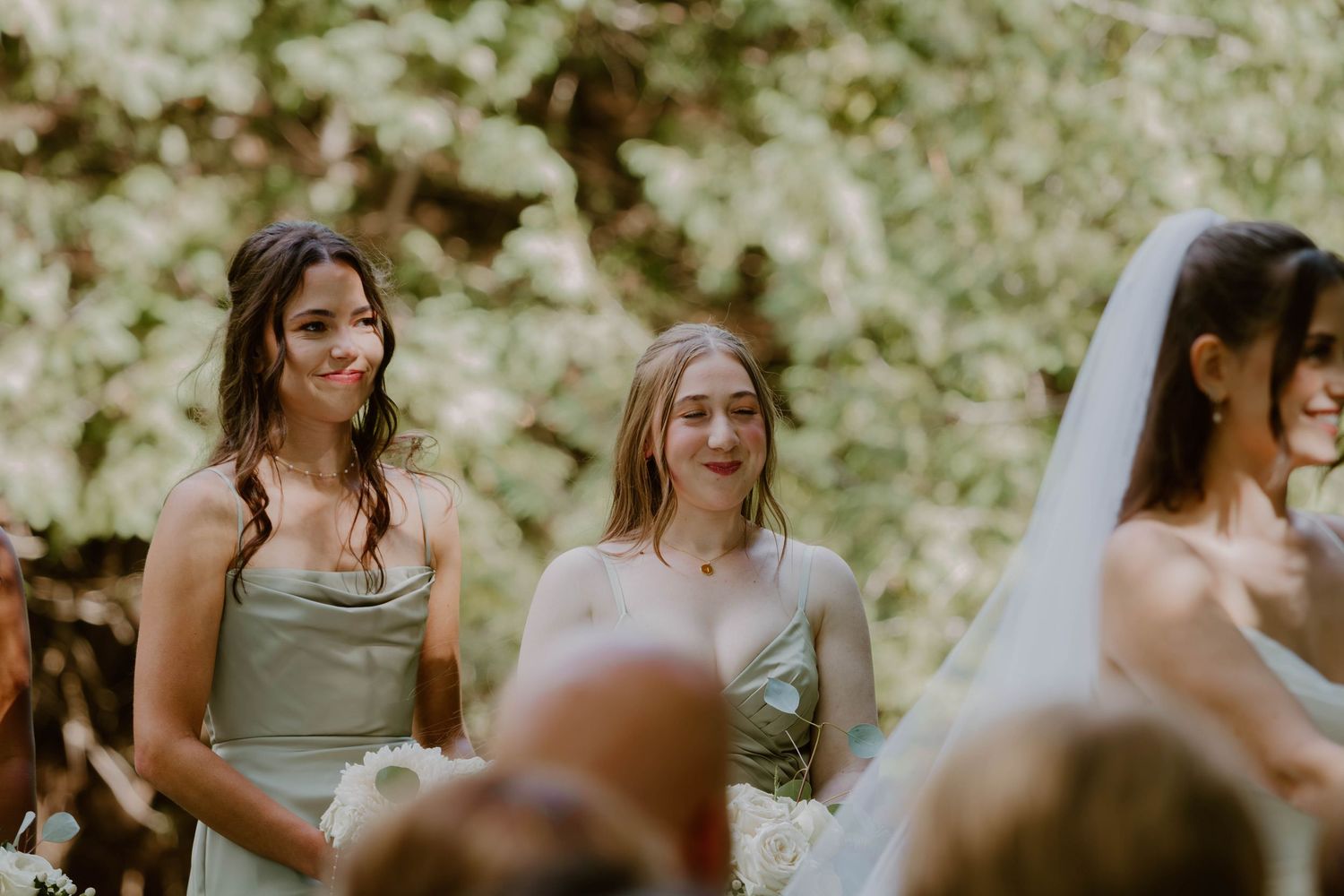 Bridesmaids in sage green dresses smile during an outdoor wedding ceremony in natural lighting.