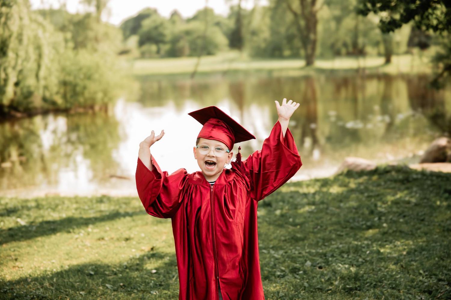 A joyful graduate in bright red regalia celebrates with raised arms by a serene pond.