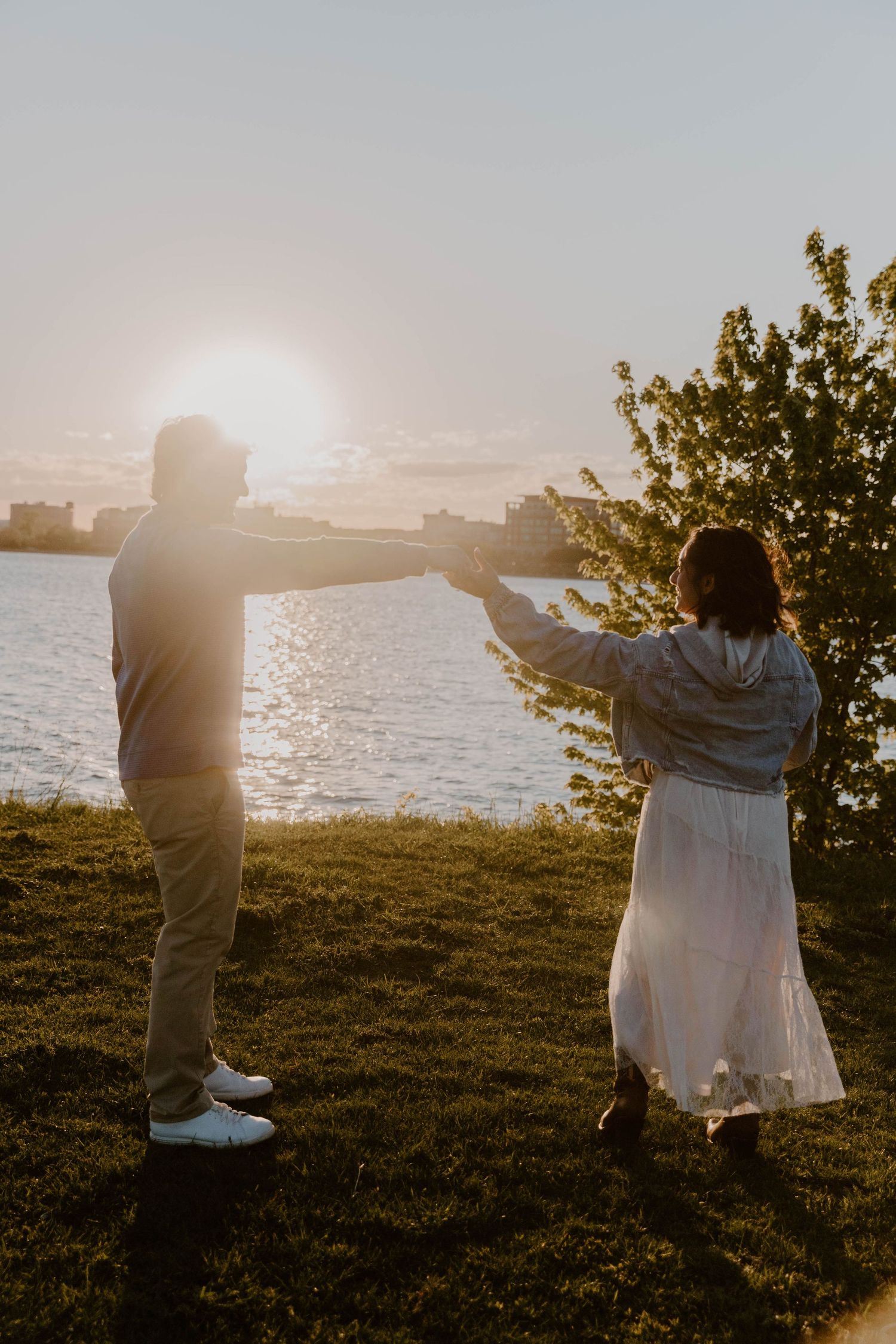 Couple holding hands at sunset by a waterfront park, woman in white dress and man in casual attire, golden hour light.
