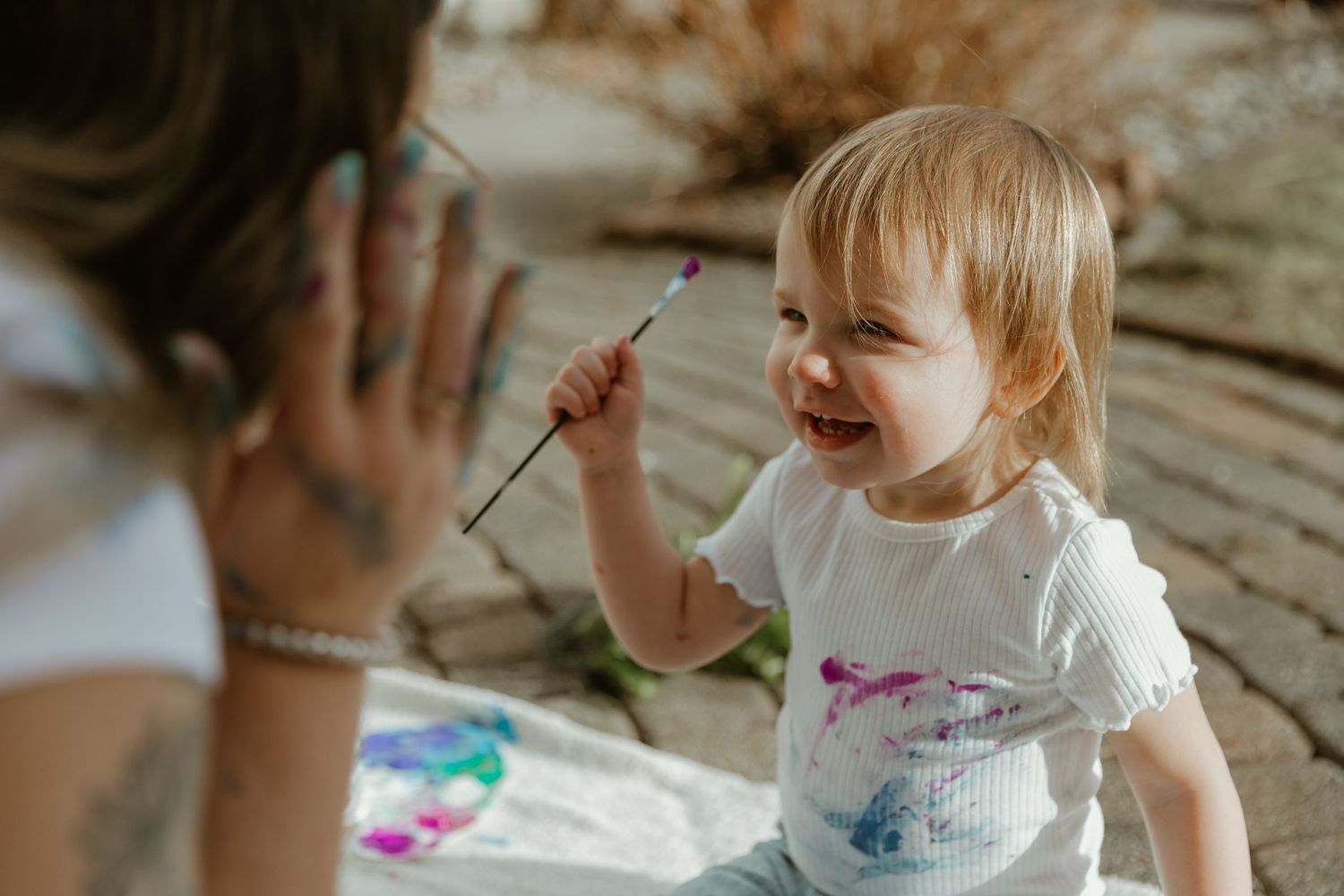 A smiling young child in a white shirt holds a small stick while sitting at a table during an art activity.