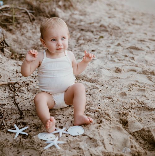 Baby in white outfit sitting on sandy beach with starfish decorations.