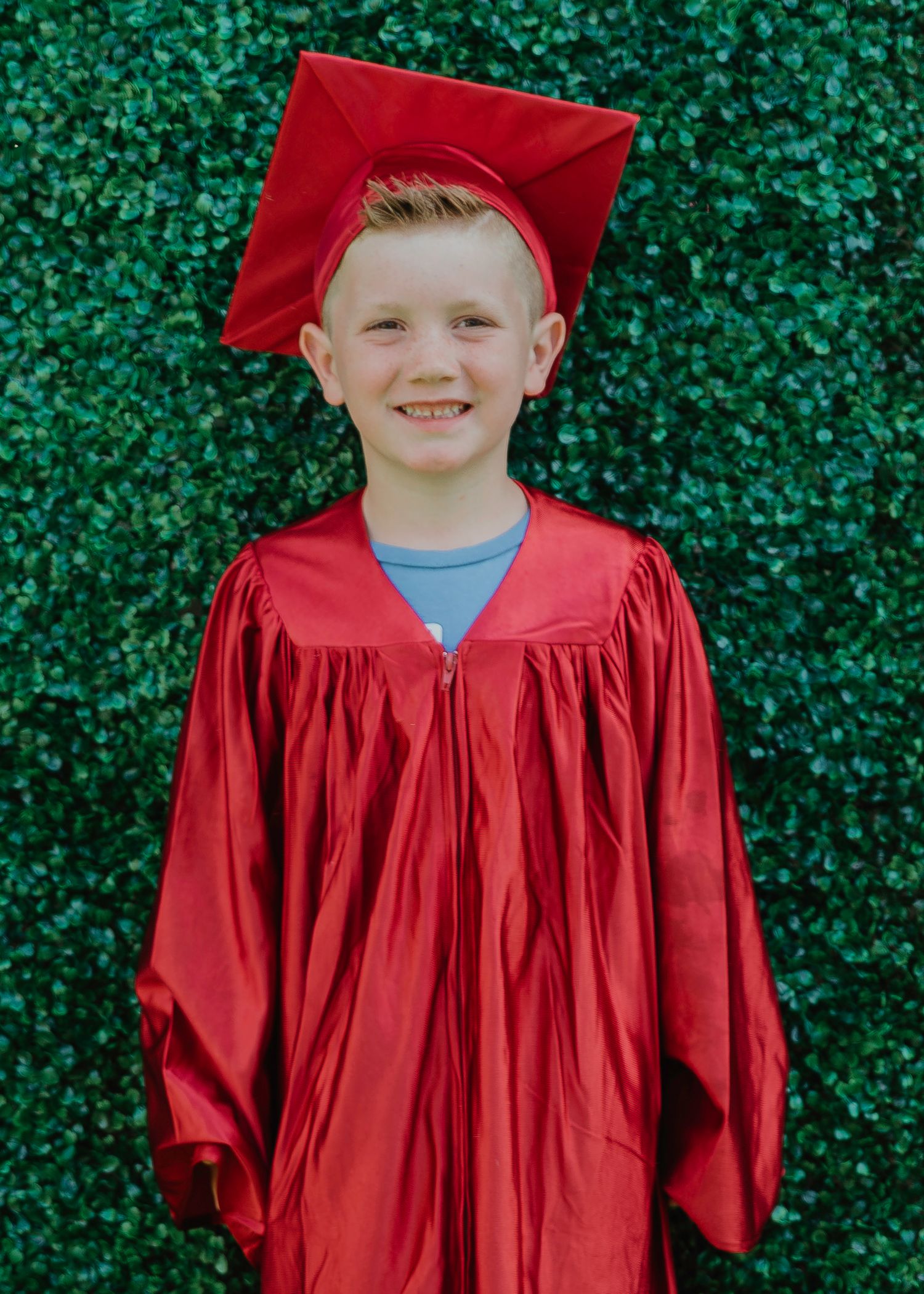 Young boy smiling in red graduation cap and gown standing in front of a green hedge wall.