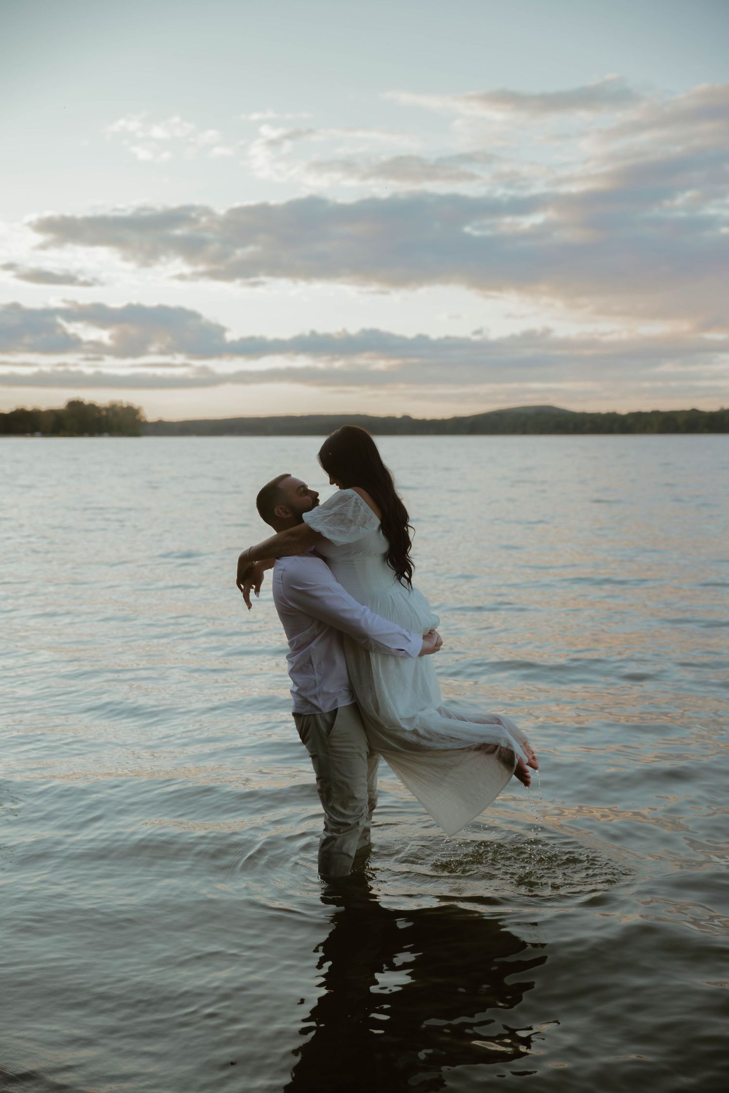 Playful couple dances in ocean water against dramatic sunset sky.