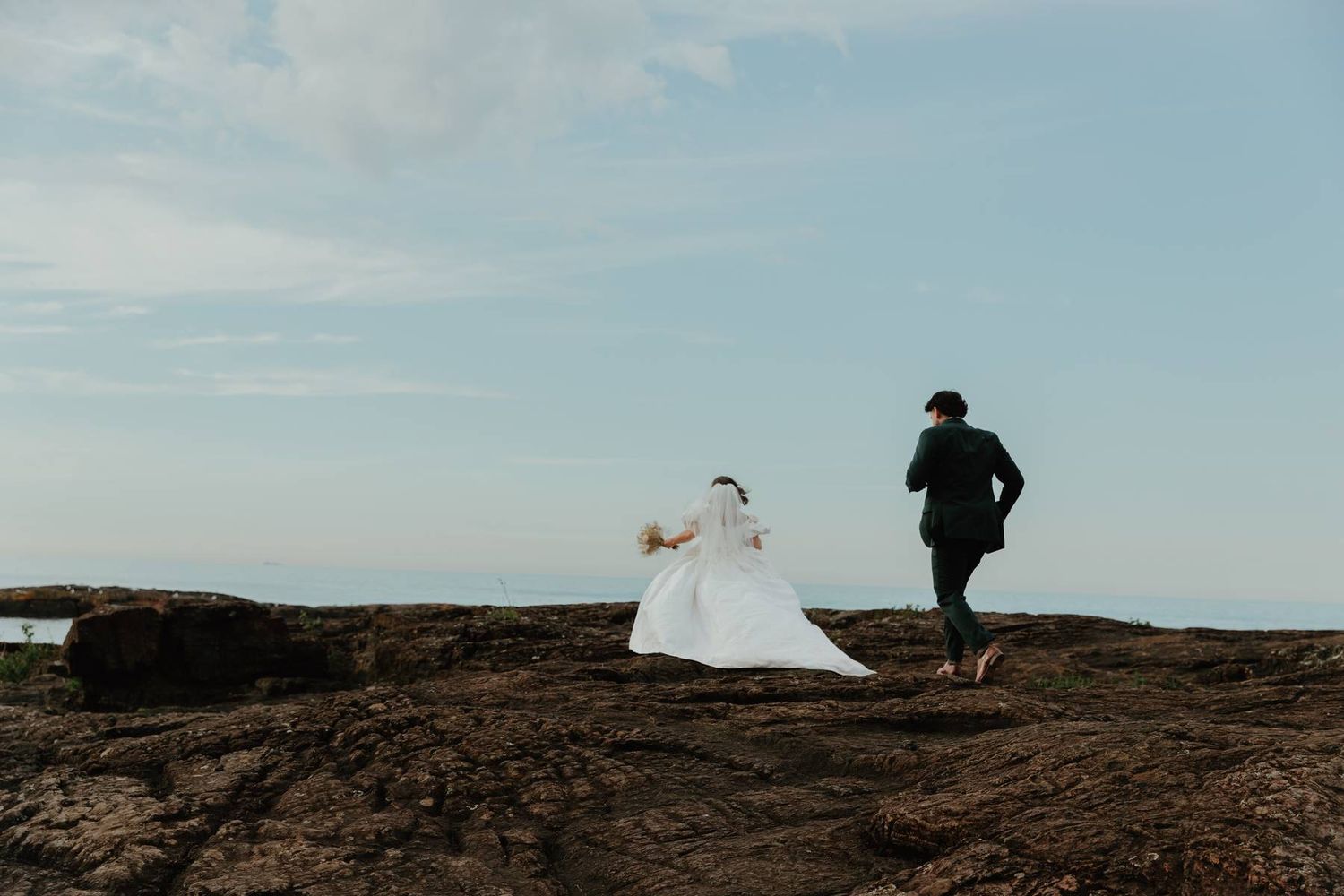 Bride in white gown and groom in black suit running on rocky coastal cliffs under a blue sky.