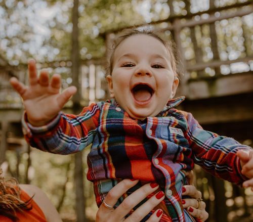 Excited baby in plaid shirt being lifted up against wooded background.