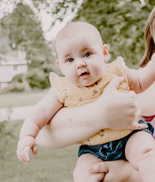 Baby wearing yellow romper held in caregiver's arms outdoors.