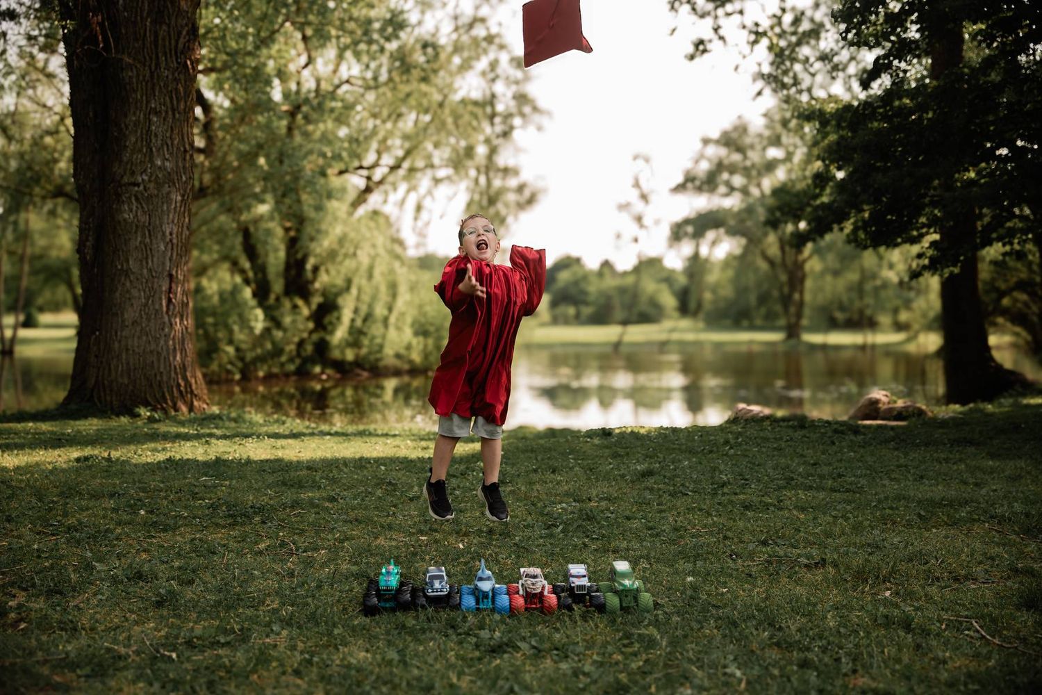 Child in red dress jumps with joy near a pond with colorful toys lined up on grass in a park setting.