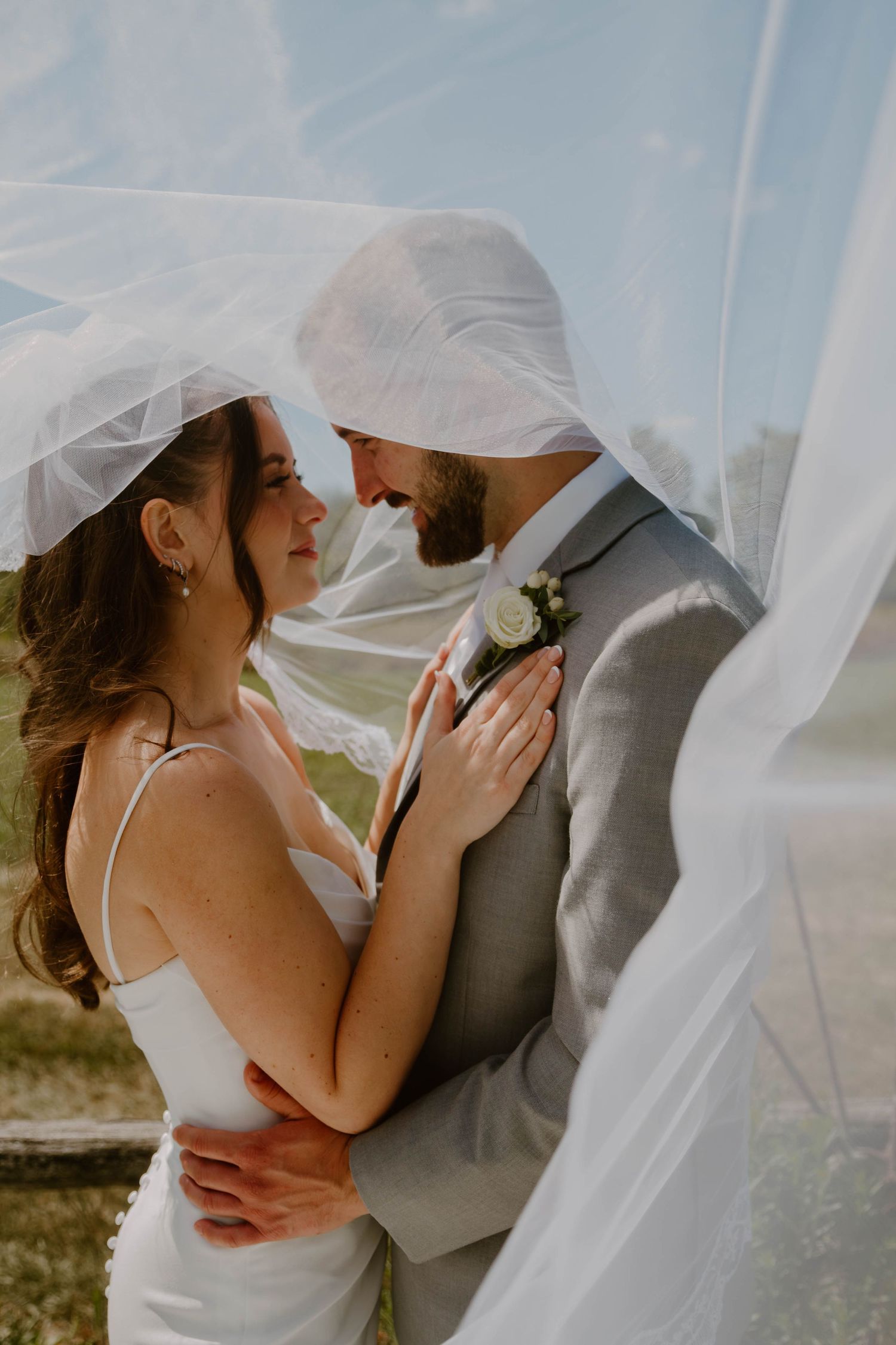 Romantic wedding couple shares an intimate moment under a flowing white veil against a natural outdoor backdrop.