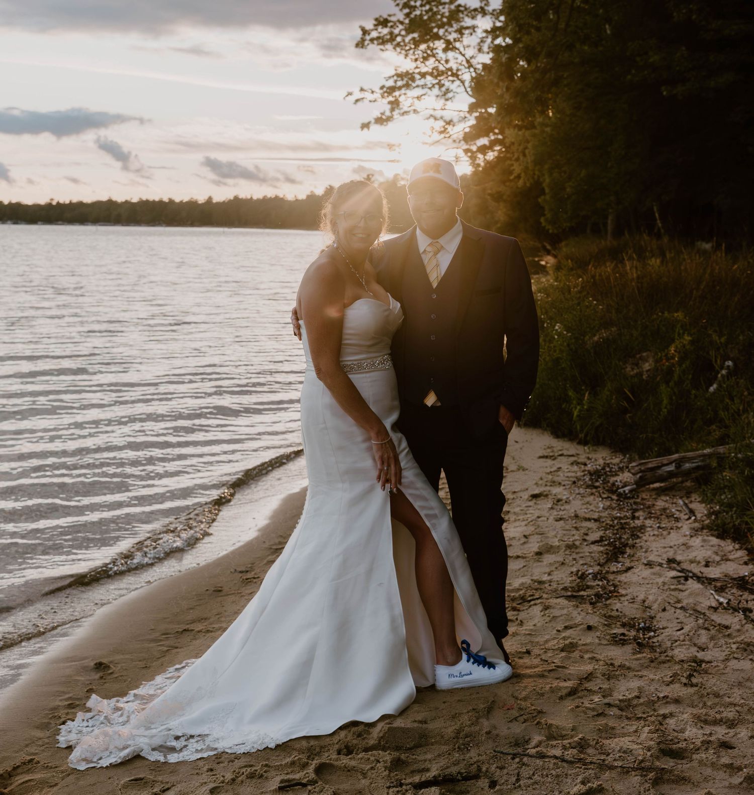 A couple shares an intimate moment on a beach at sunset with water and trees in the background.