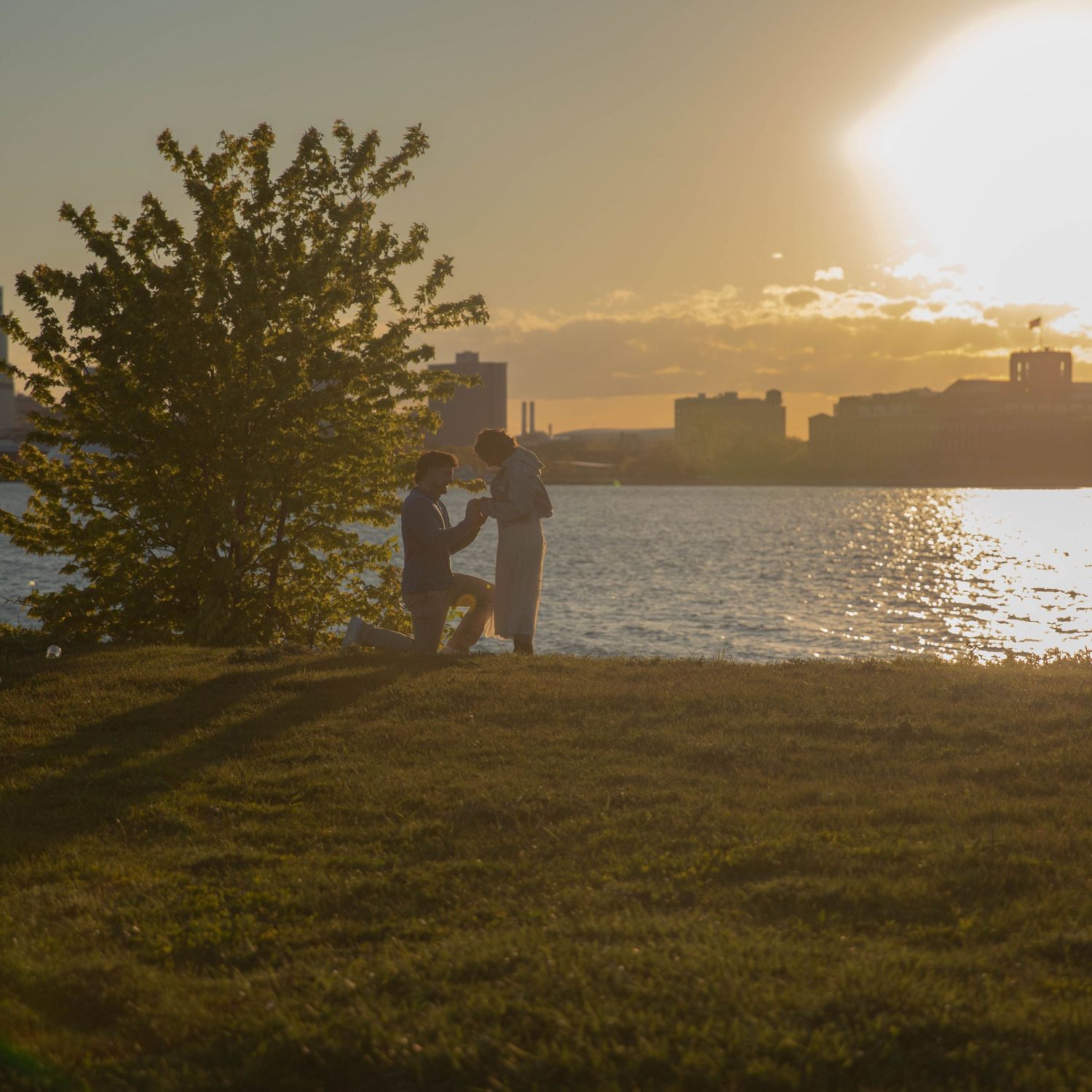 Couple sharing a romantic proposal moment on a grassy riverbank at golden sunset with city skyline backdrop.