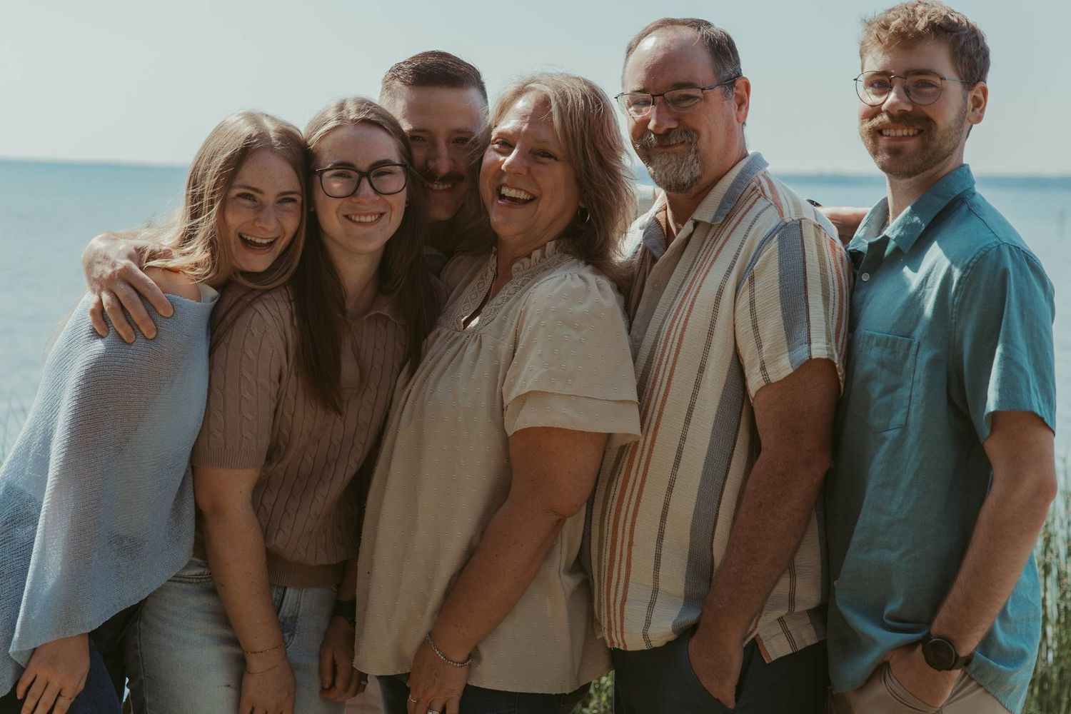 Six smiling family members pose together outdoors near a lake, dressed casually in a warm, joyful group portrait.