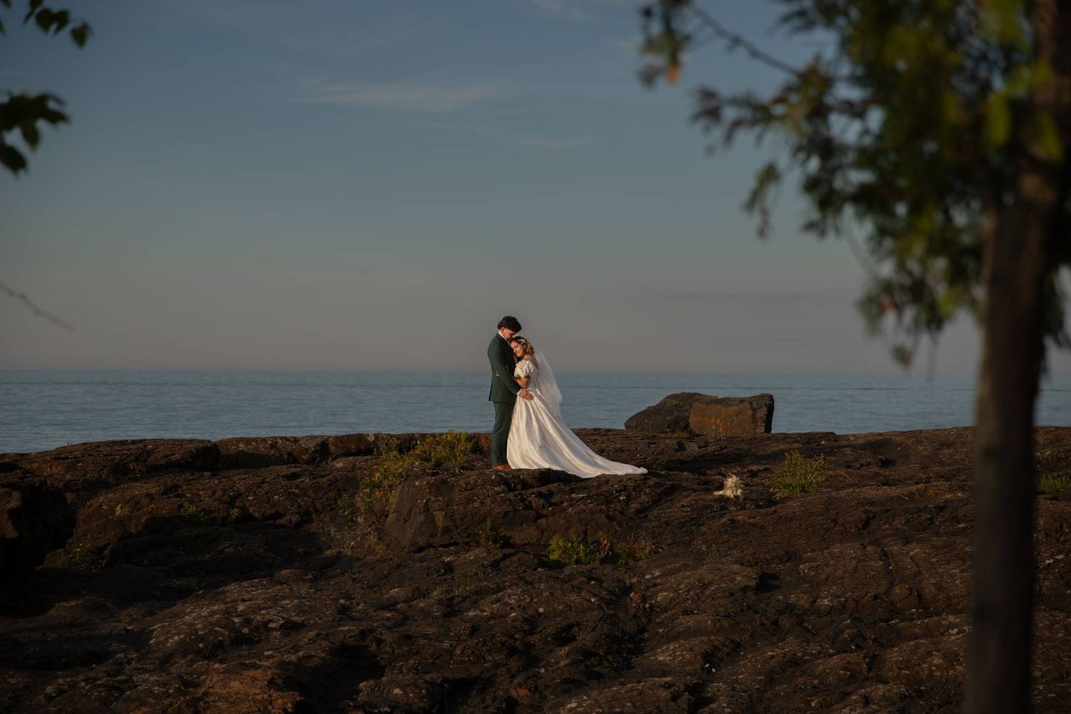 Bride and groom embrace on rocky coastal cliffs with ocean horizon backdrop during golden hour wedding portrait.