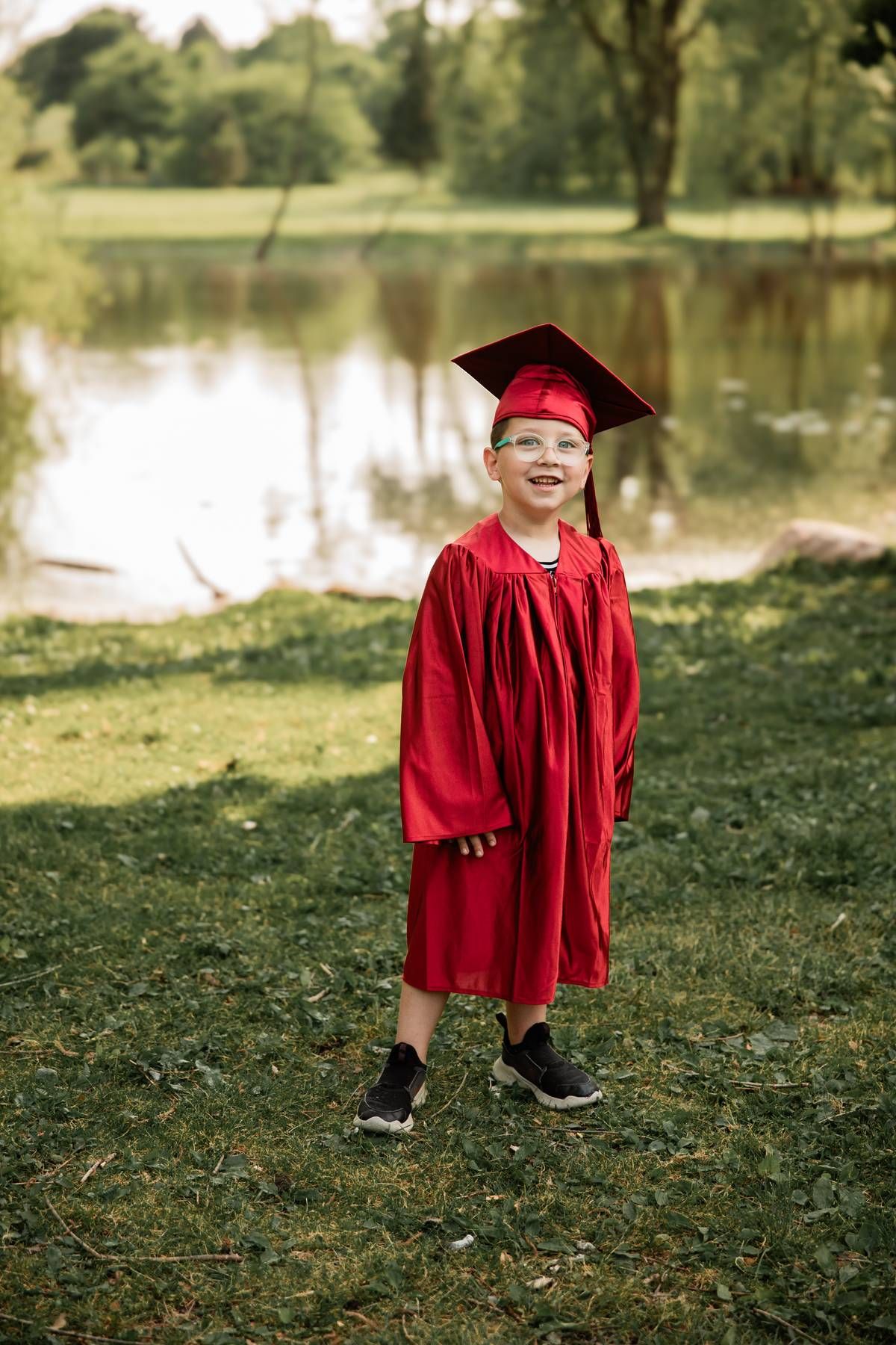 A small graduate wearing a bright red cap and gown stands near a pond on a grassy lawn.