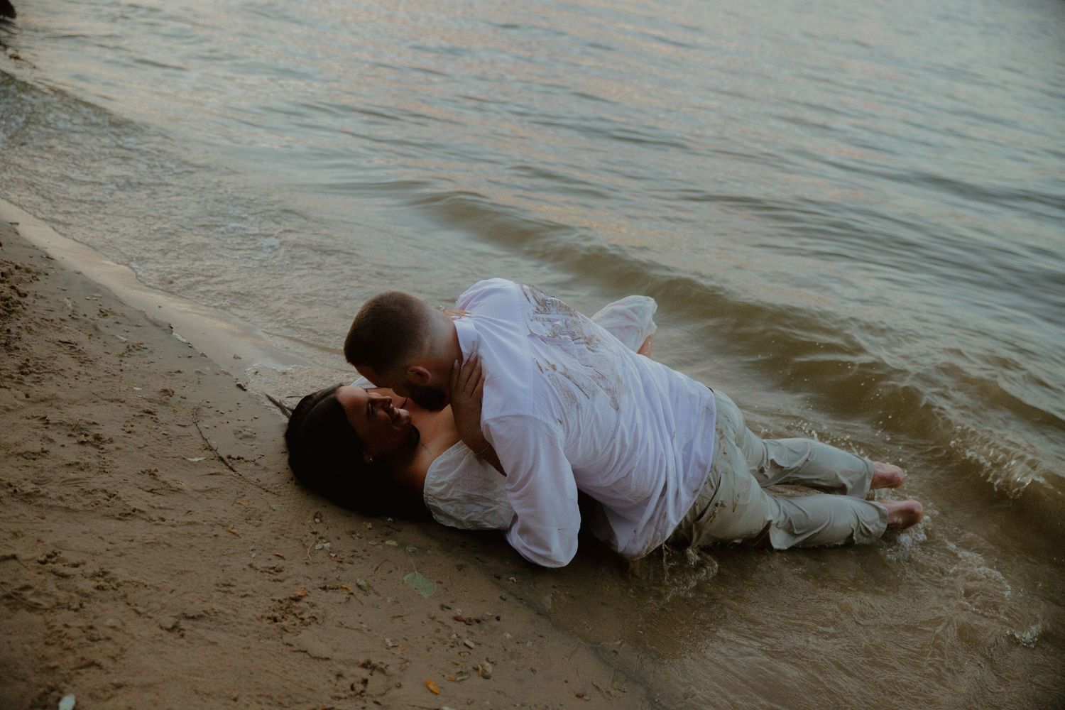 A couple in white clothing lying together on a sandy beach at sunset.
