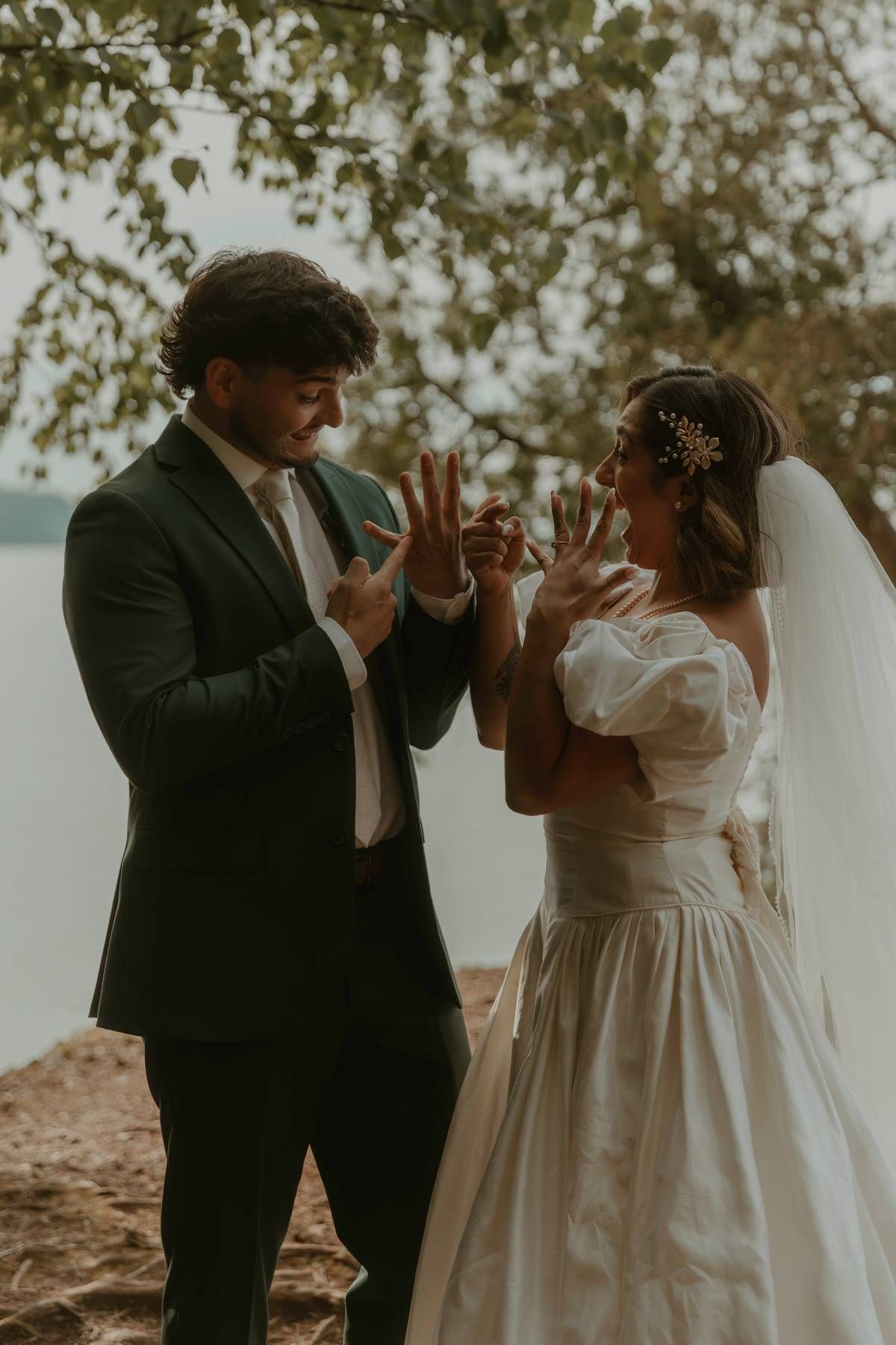 Bride and groom exchanging rings outdoors near a lake, bride in white gown with veil, groom in dark suit.