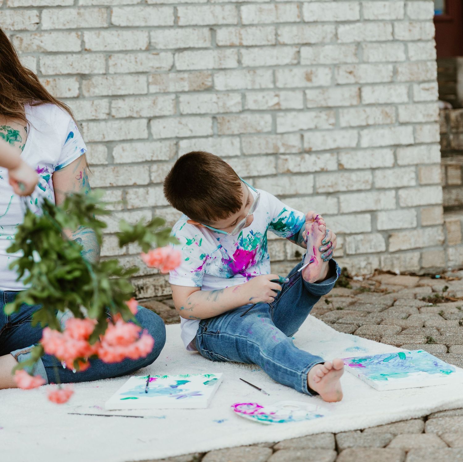 A creative art session leaves two people covered in vibrant paint splatters while sitting on a white blanket near a brick wall.
