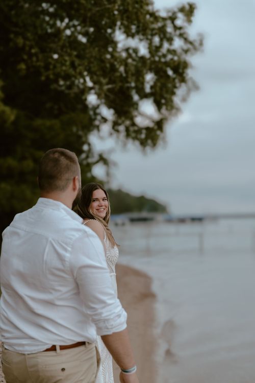 A couple in white clothing sharing a romantic moment on a beach at dusk with boats visible in the background.