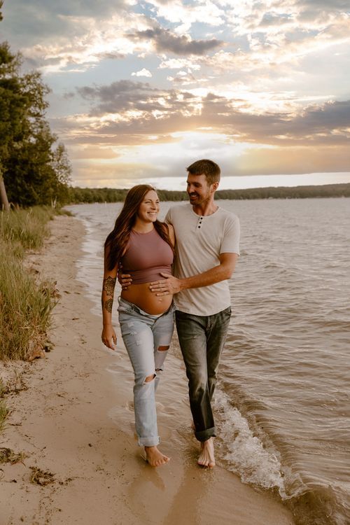 Expectant couple walking together on beach shoreline during golden hour sunset.
