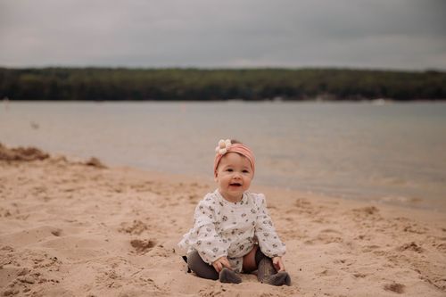 A small child in a floral outfit sits on a sandy beach near a peaceful lake.