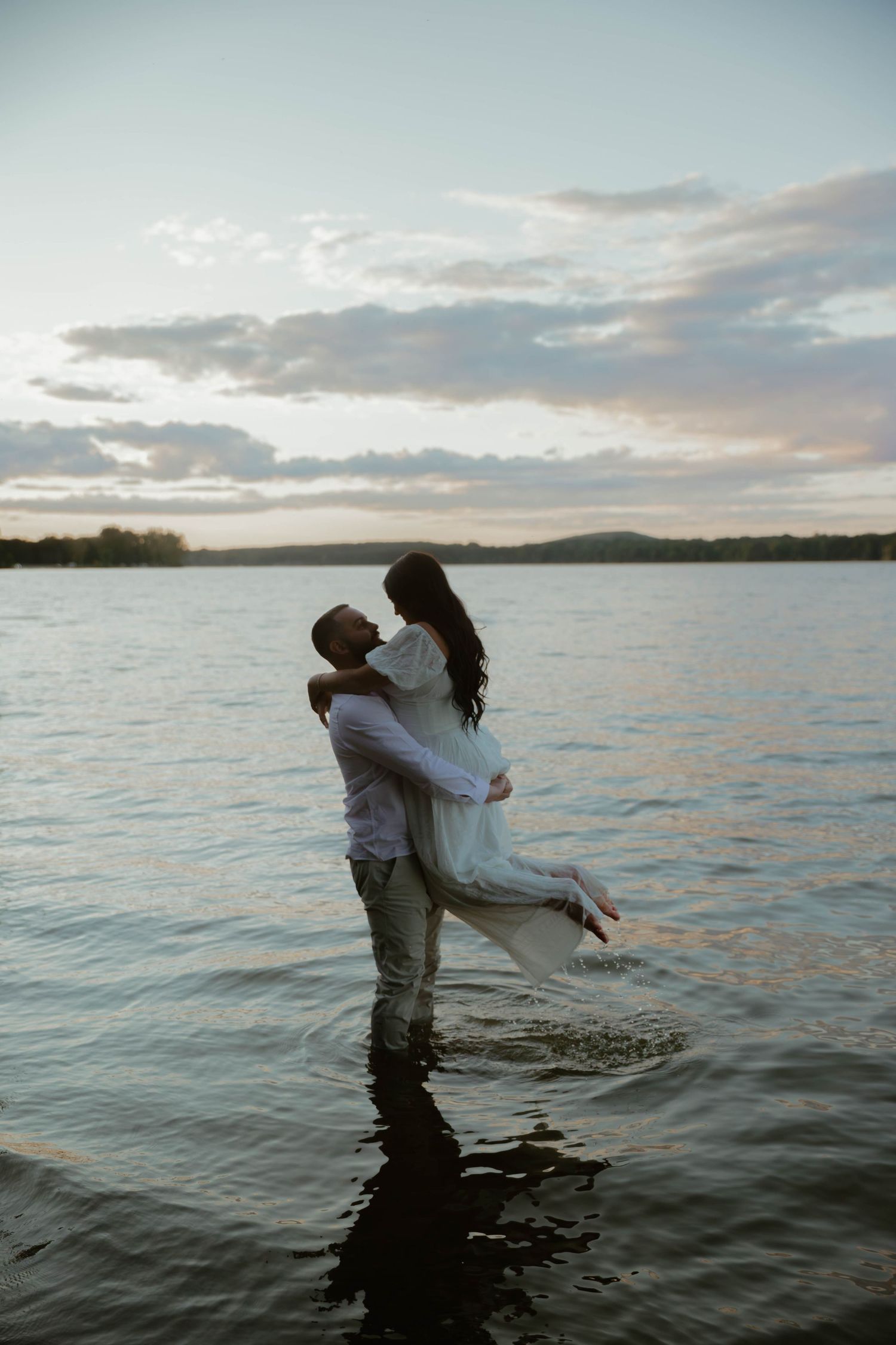 A couple embraces while standing in shallow water at sunset, creating a romantic silhouette against the lake and sky.