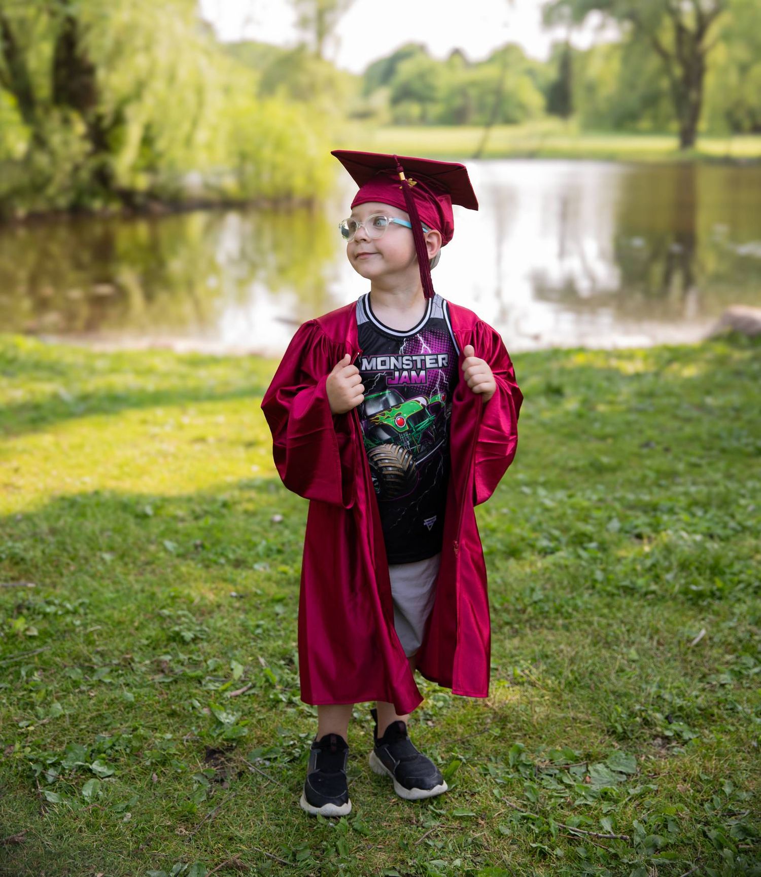 A graduate poses confidently in red commencement regalia and casual outfit by a lakeside setting.