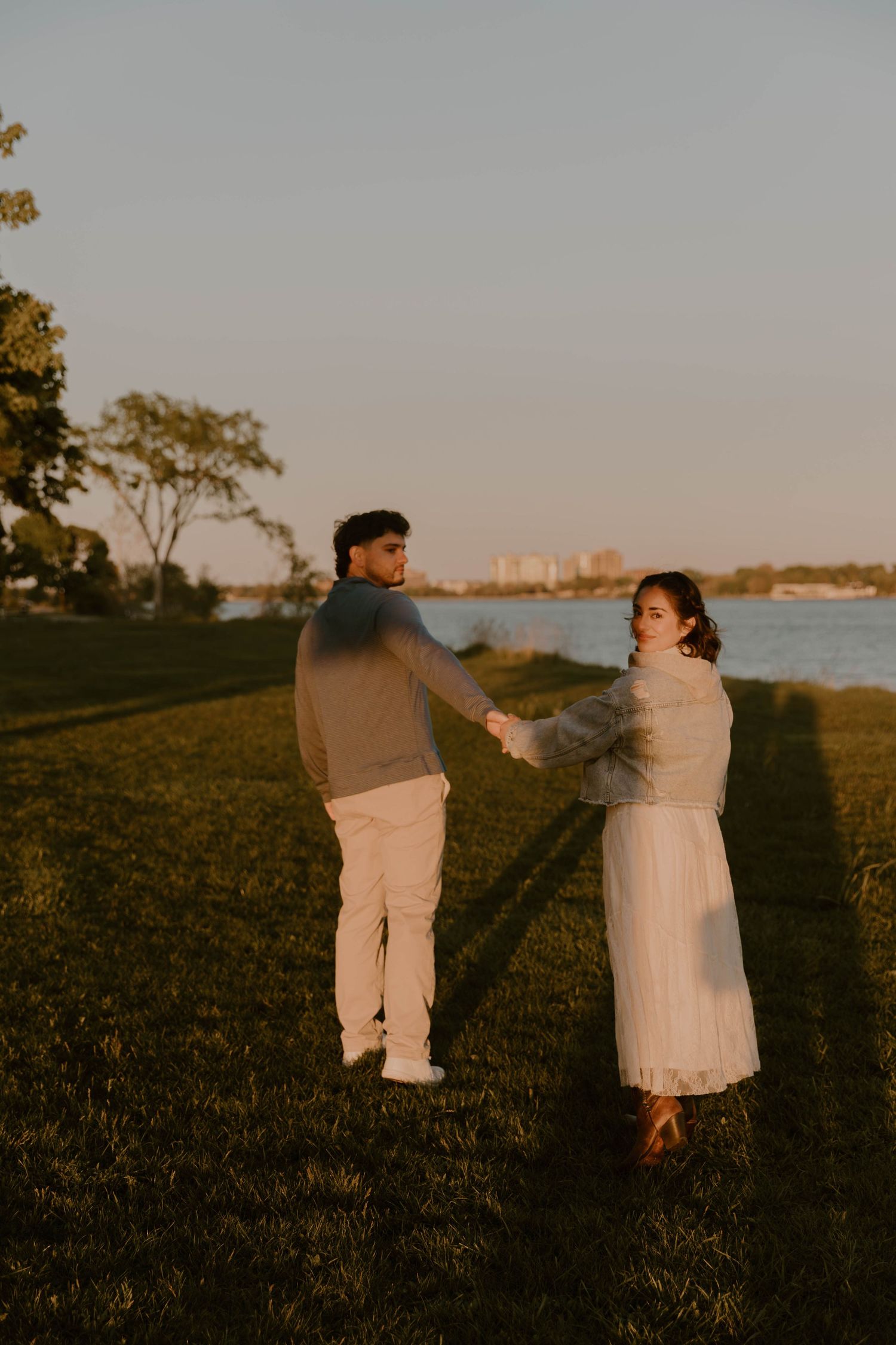Couple holding hands at golden hour by a waterfront park, woman in white dress looking back over her shoulder.