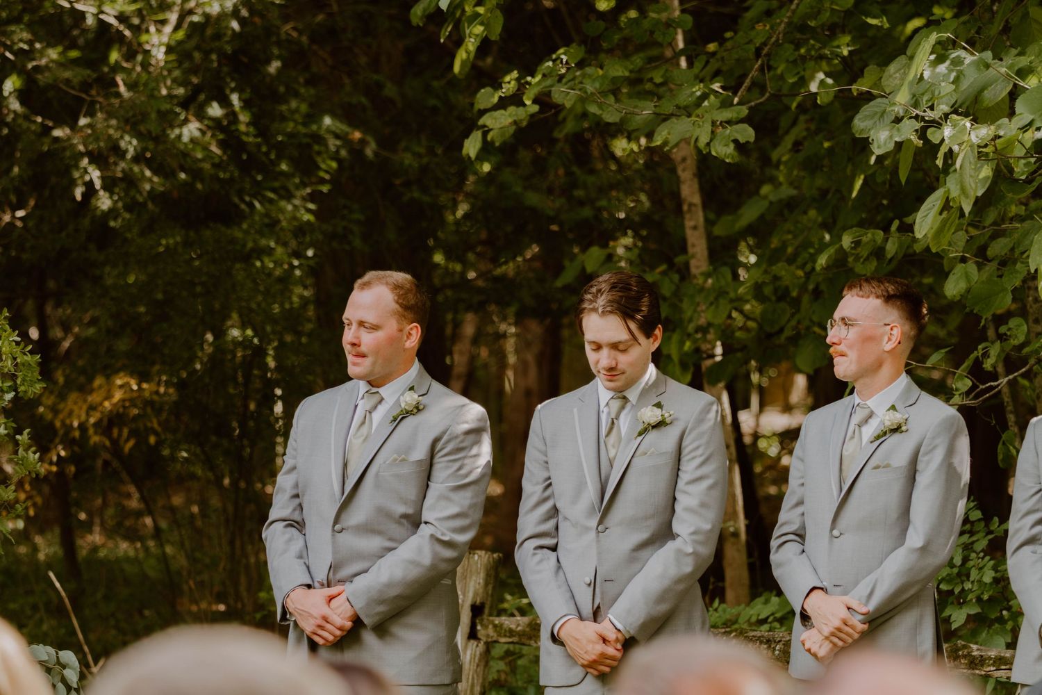Groomsmen in matching light gray suits stand in line during an outdoor wedding ceremony.