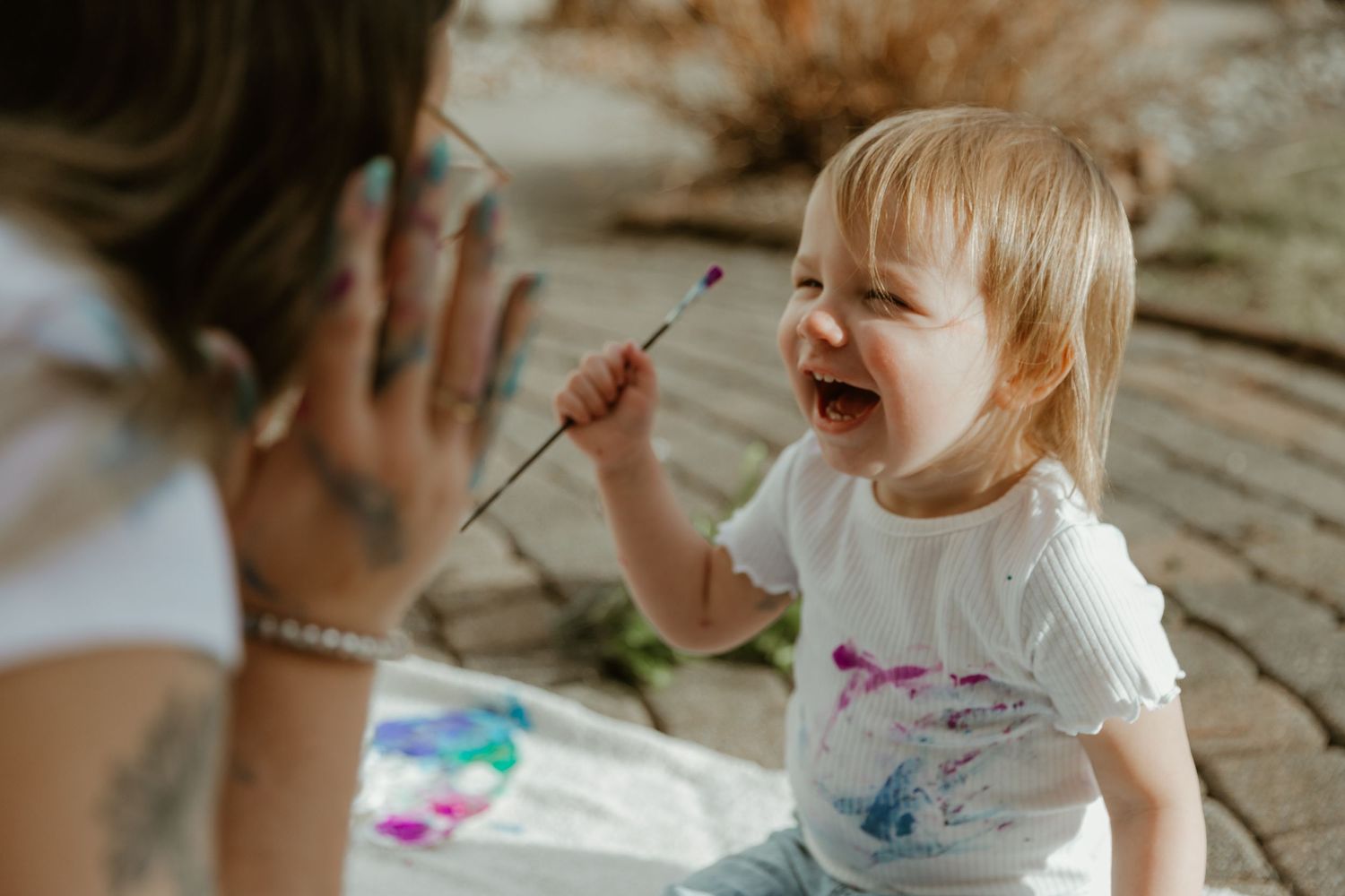 A joyful toddler in a white t-shirt plays and laughs during a creative activity outdoors.