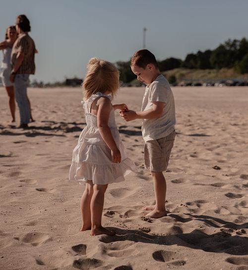 Two children in light colored clothing interacting on a sandy beach.
