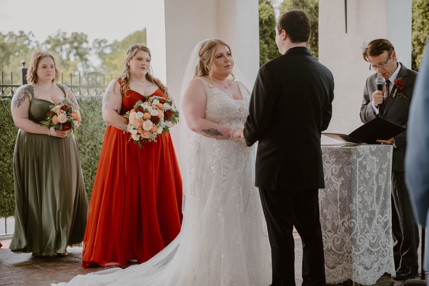 Wedding party stands together during outdoor ceremony with bridesmaids in red and olive green dresses.