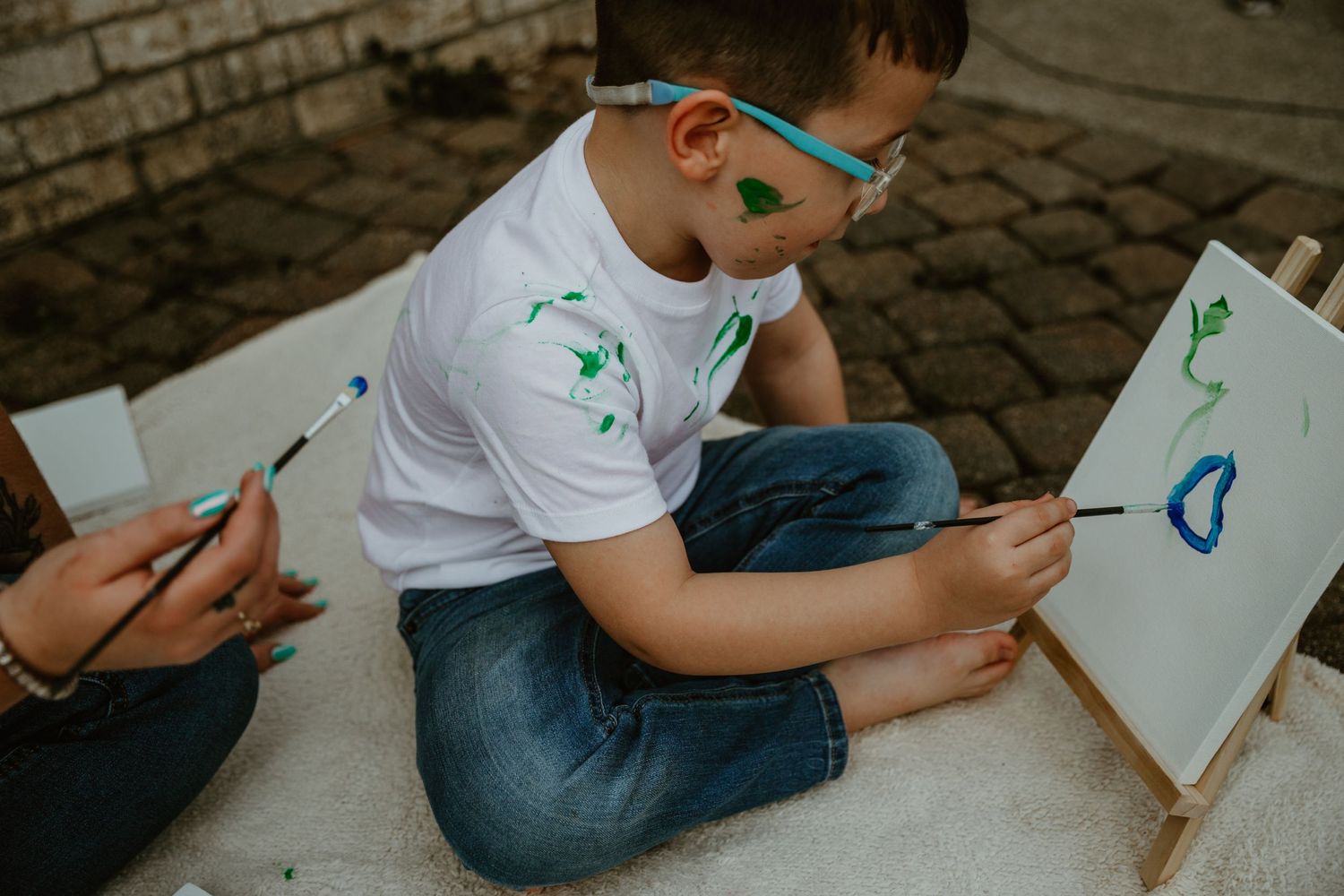 Two children sitting on pavement drawing with blue markers on white sketchbooks during an outdoor art activity.