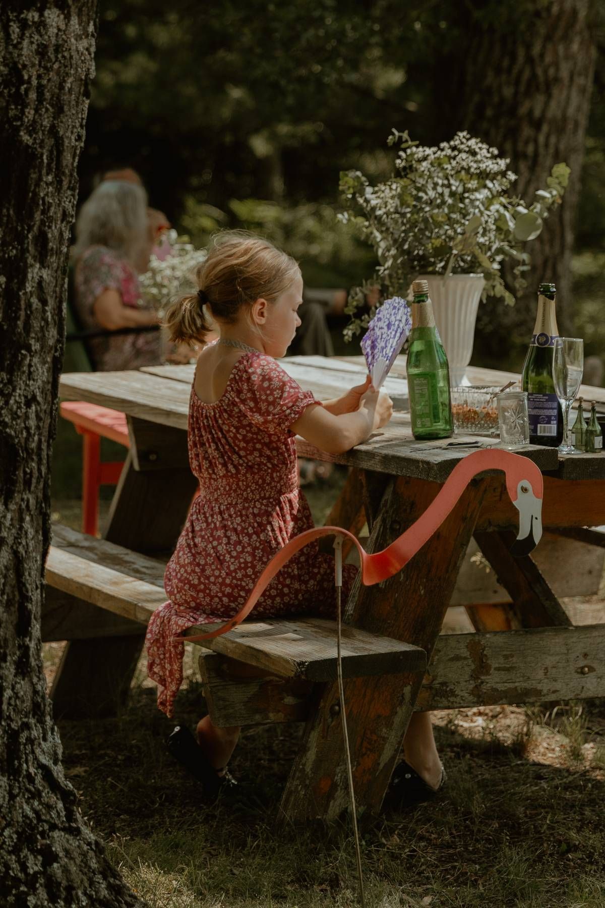 Young girl in floral dress sitting at outdoor picnic table with flowers and bottles in a forest setting.