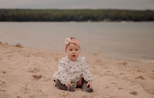 A small child in floral clothing sits on a sandy beach by a tranquil lake.