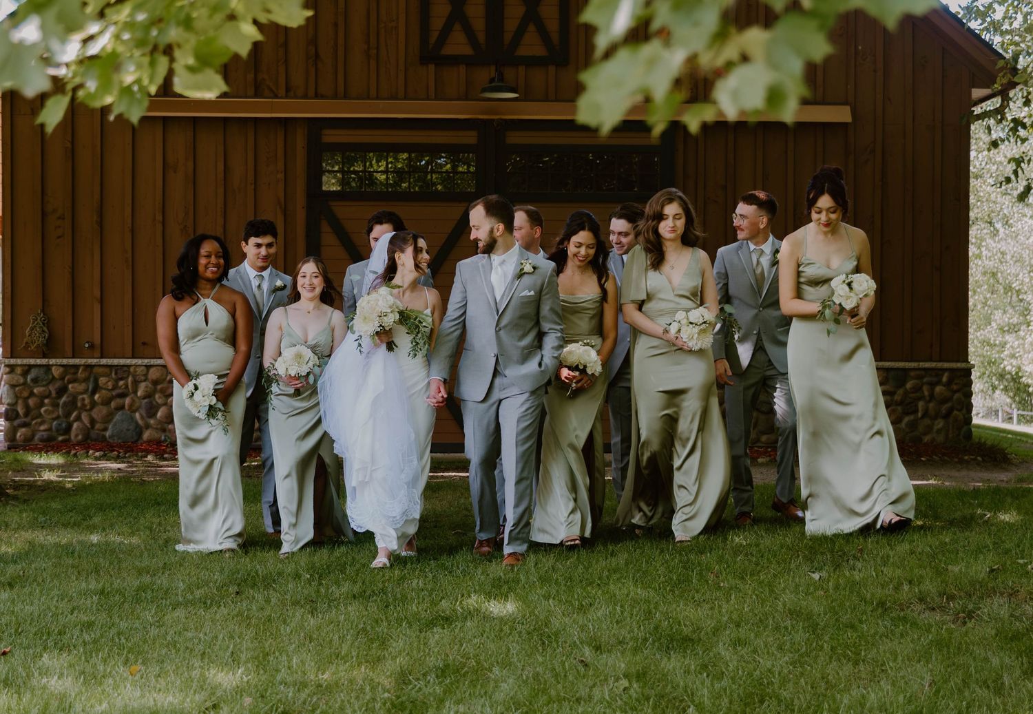 Wedding party in light gray and white attire poses together in front of rustic brown barn exterior.