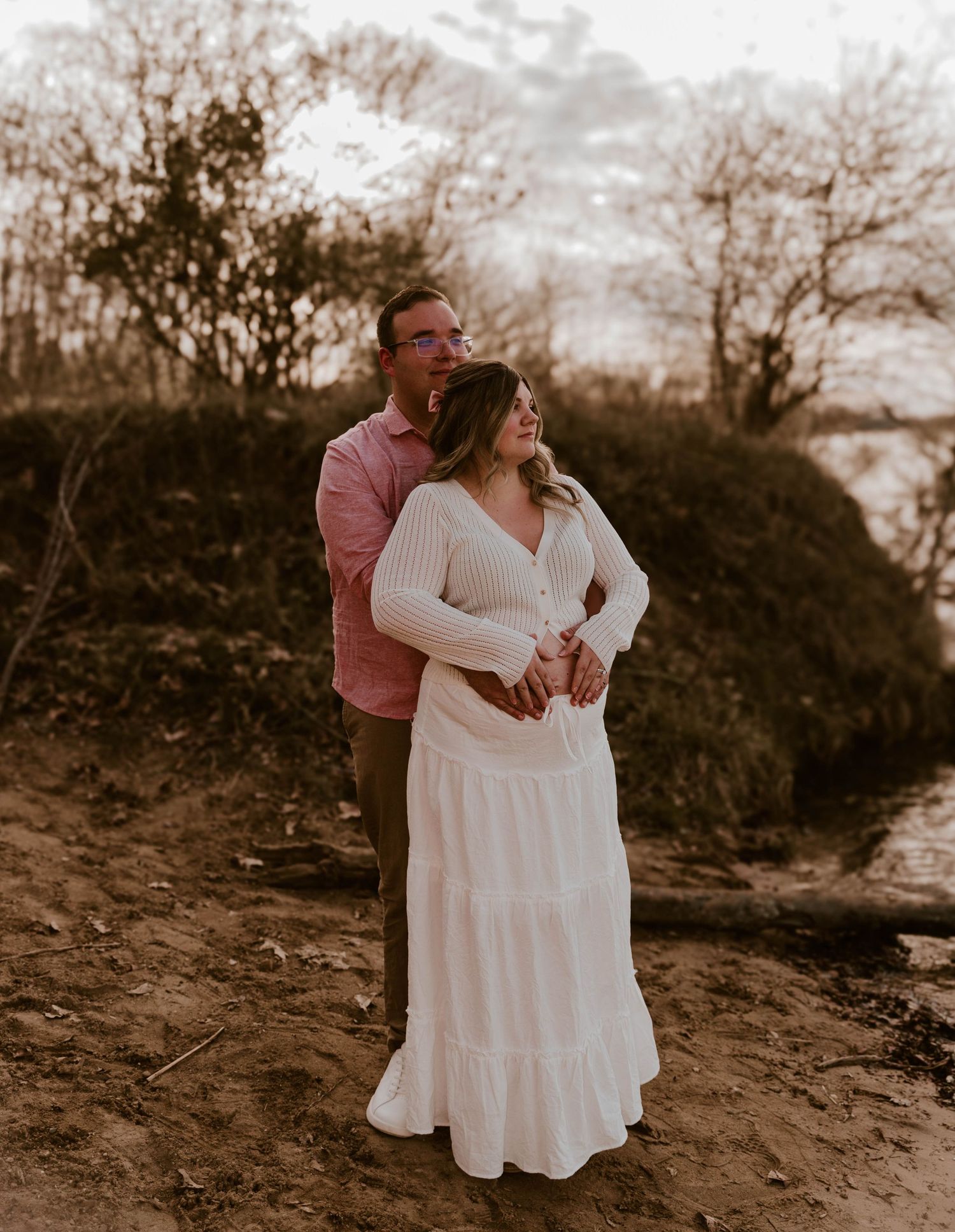 Couple embraces in romantic sunset moment on dirt path near water with moody warm lighting and natural scenery.