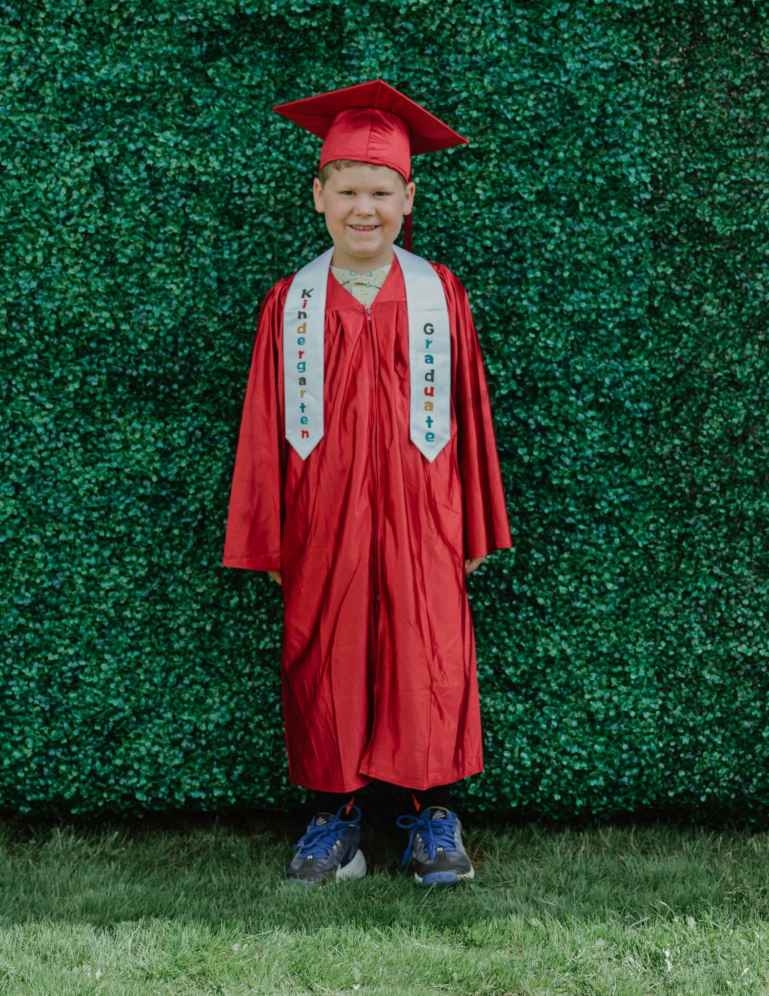 Young boy in red graduation cap and gown with white stoles smiling in front of green hedge wall.