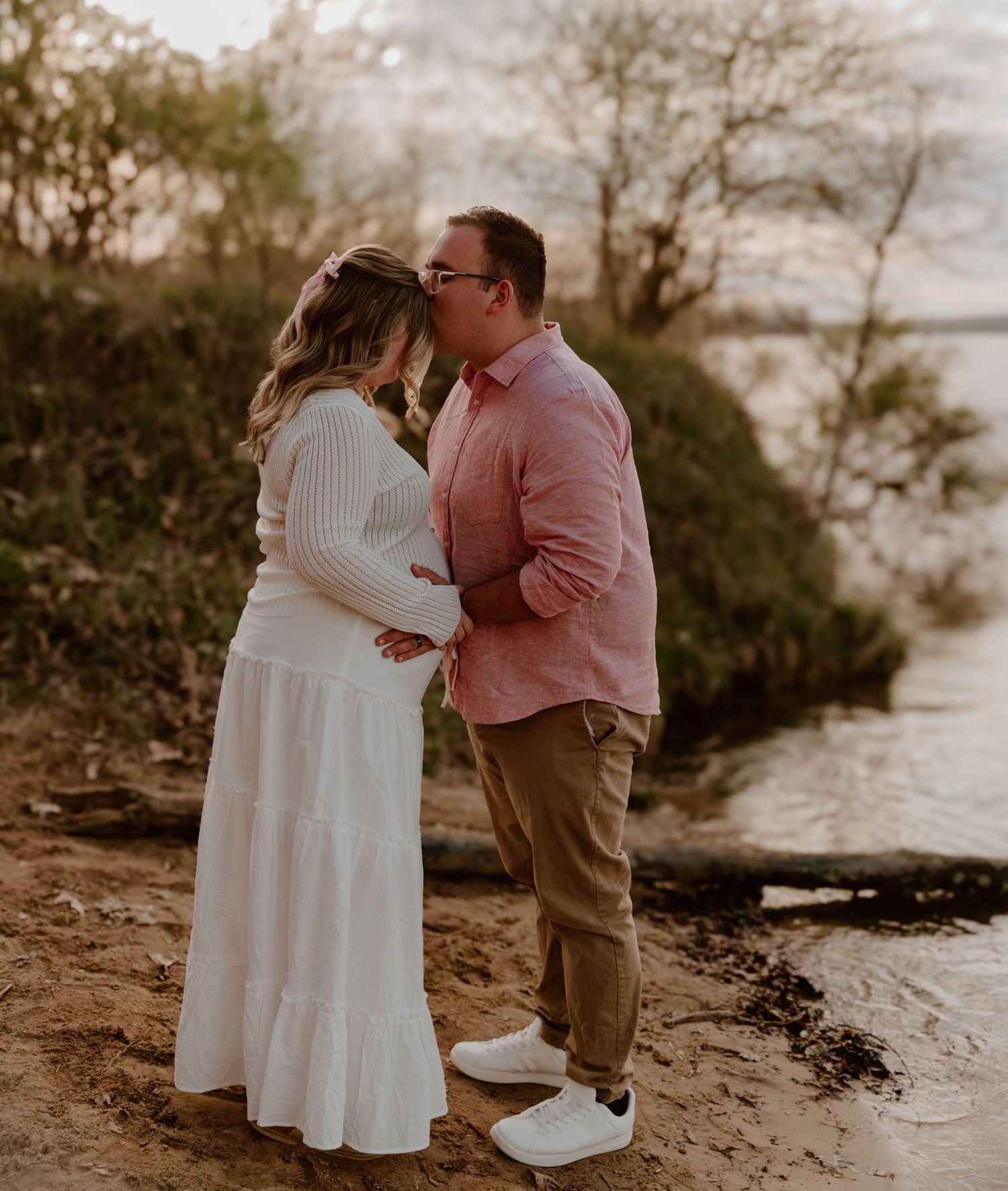 A couple in romantic pose standing by waterside at sunset, with person in white dress showing pregnancy.