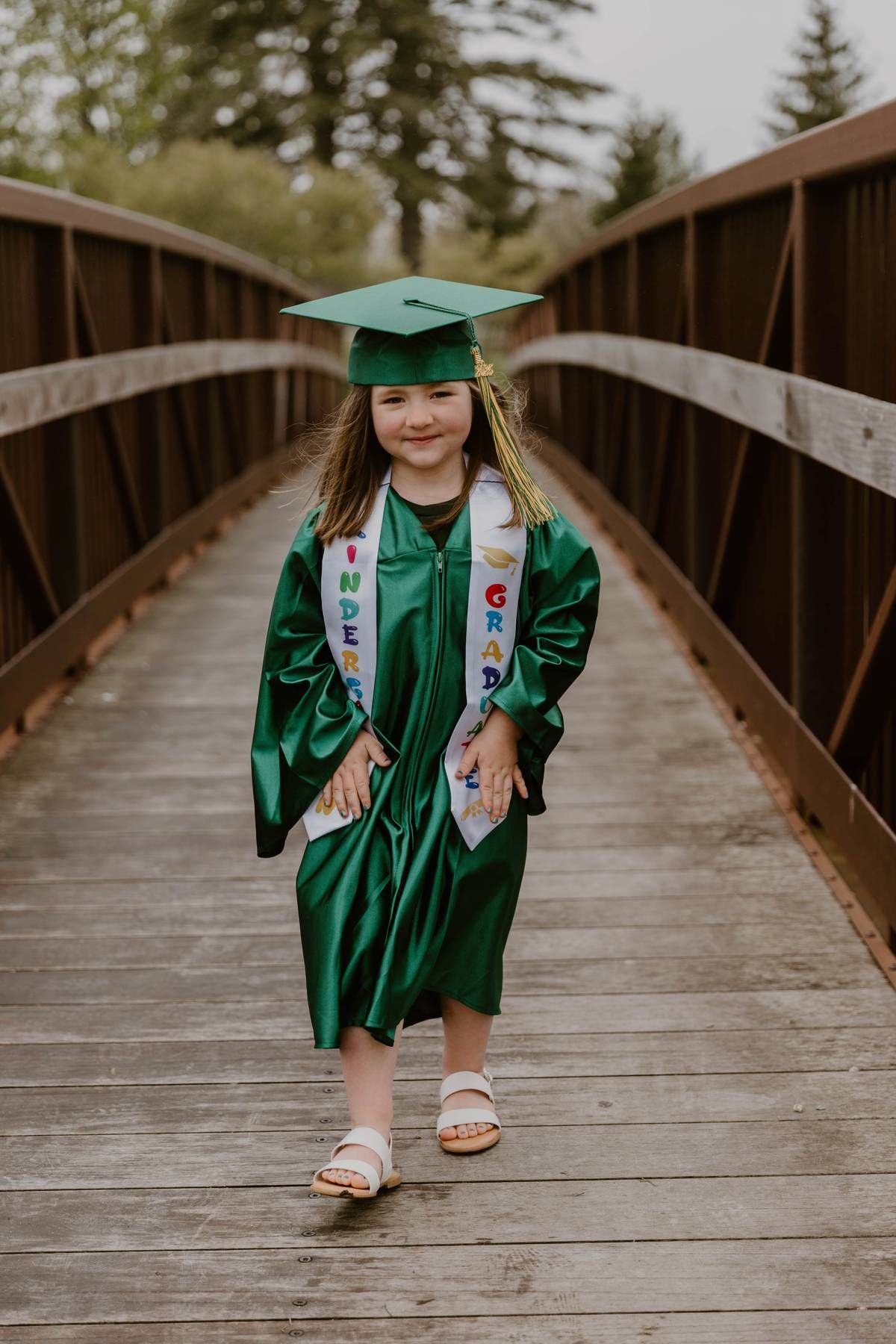 Graduate in green cap and gown walks across wooden bridge in outdoor setting during graduation photo session.