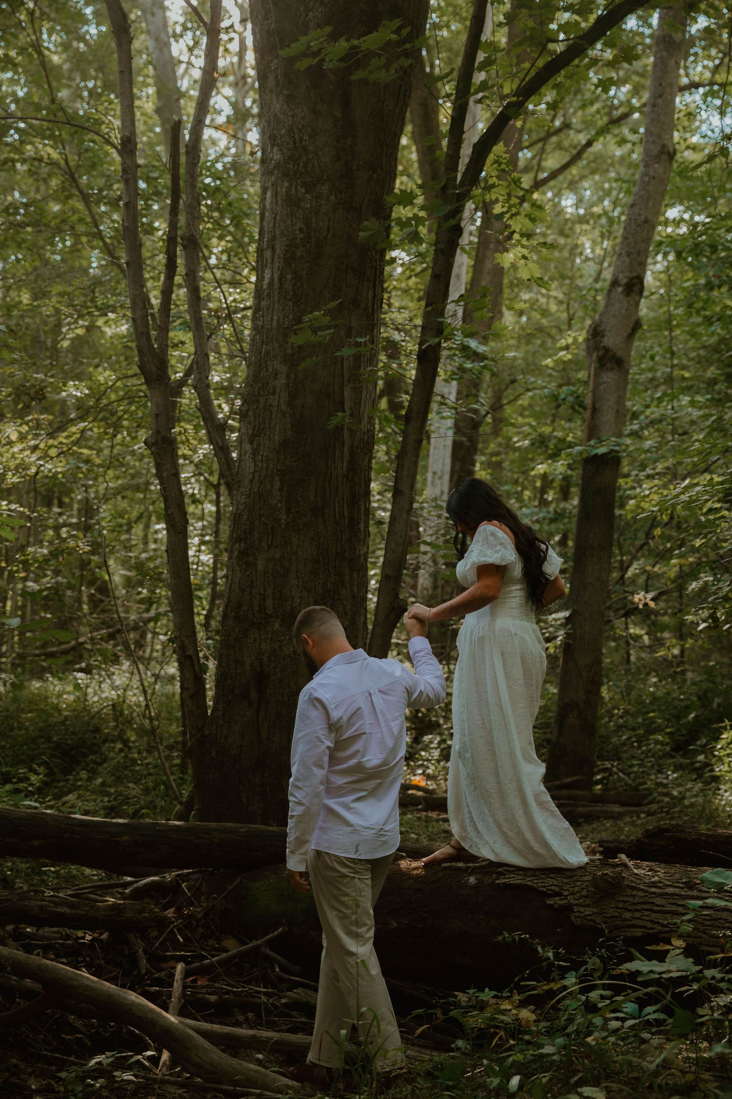 People in white clothing walk through a serene forest setting with tall trees and dappled sunlight.