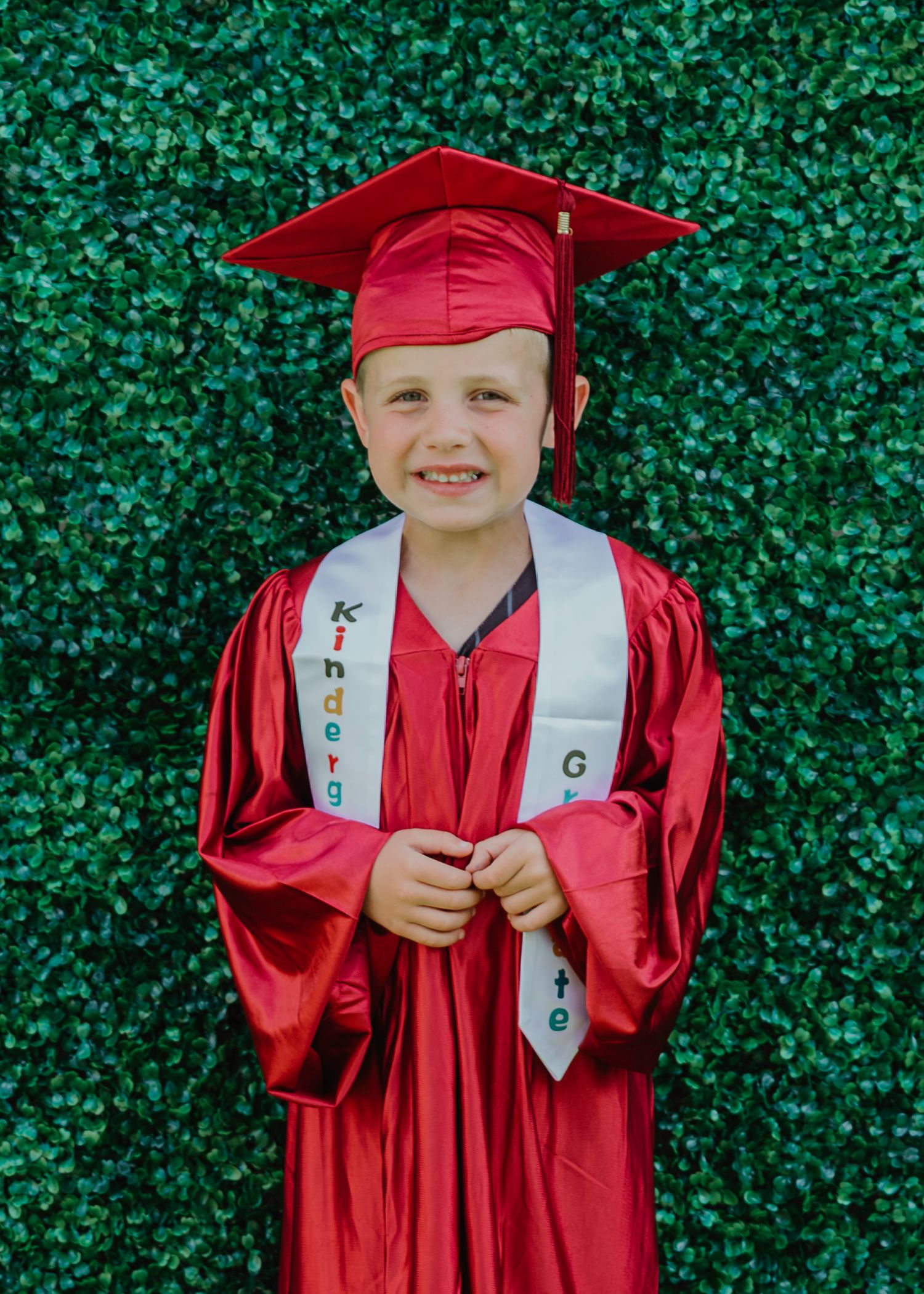 Young child in red kindergarten graduation cap and gown smiling in front of green ivy wall.