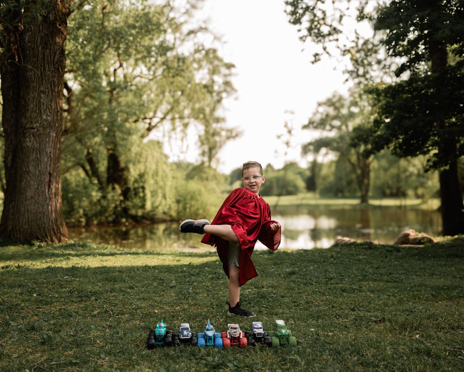 Child in a red dress balances on one leg above colorful toys lined up on grass near a pond.