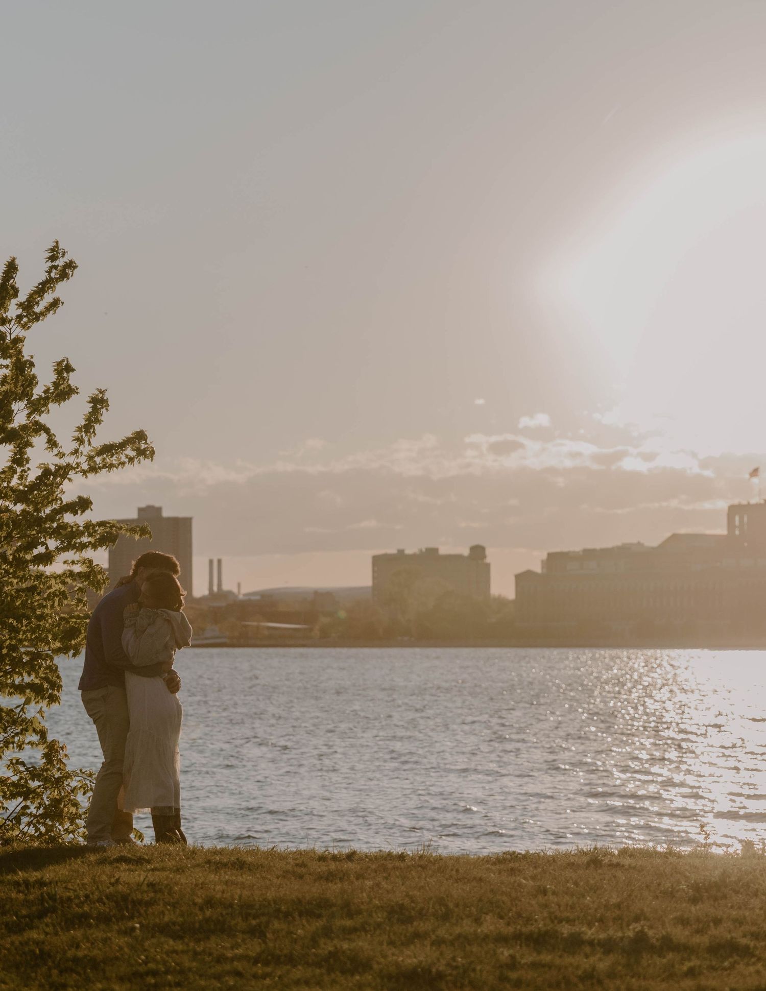 Couple embracing at golden hour by a waterfront with a city skyline in the background.