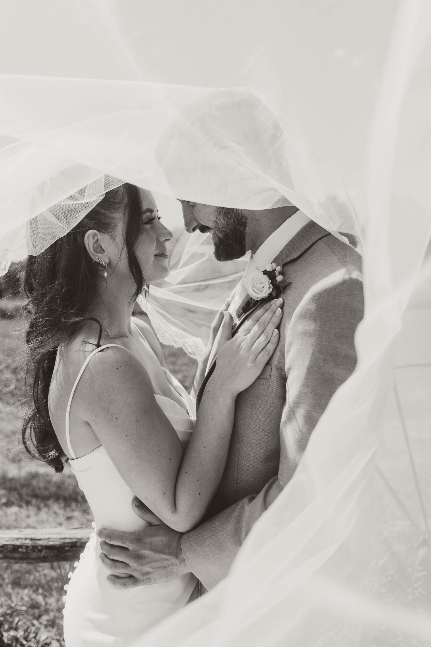 A romantic black and white wedding photo of a couple embracing under a flowing veil in an intimate moment.