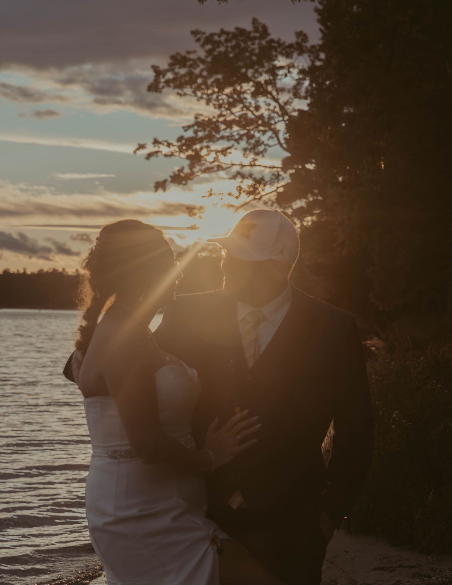 A couple embraces by a lake at sunset with silhouetted trees and glowing golden light in the background.