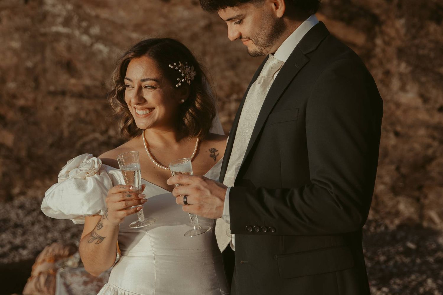 Smiling bride and groom toast with champagne glasses in a warmly lit rustic stone setting.