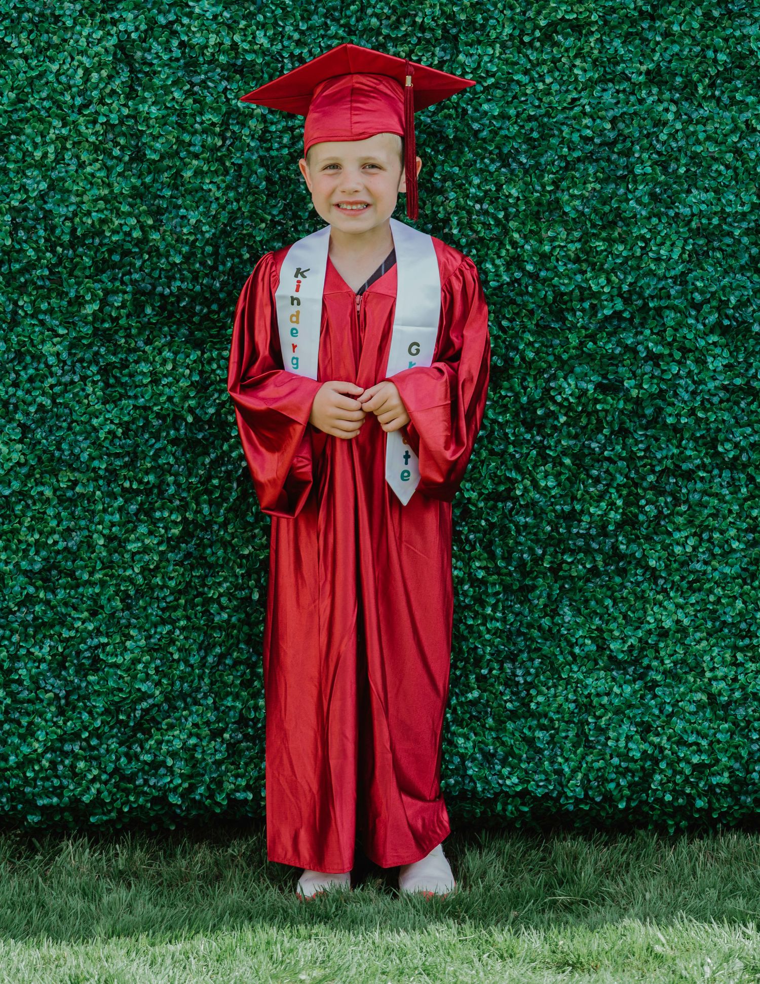 Young child smiling in red kindergarten graduation cap and gown, standing in front of lush green hedge wall.