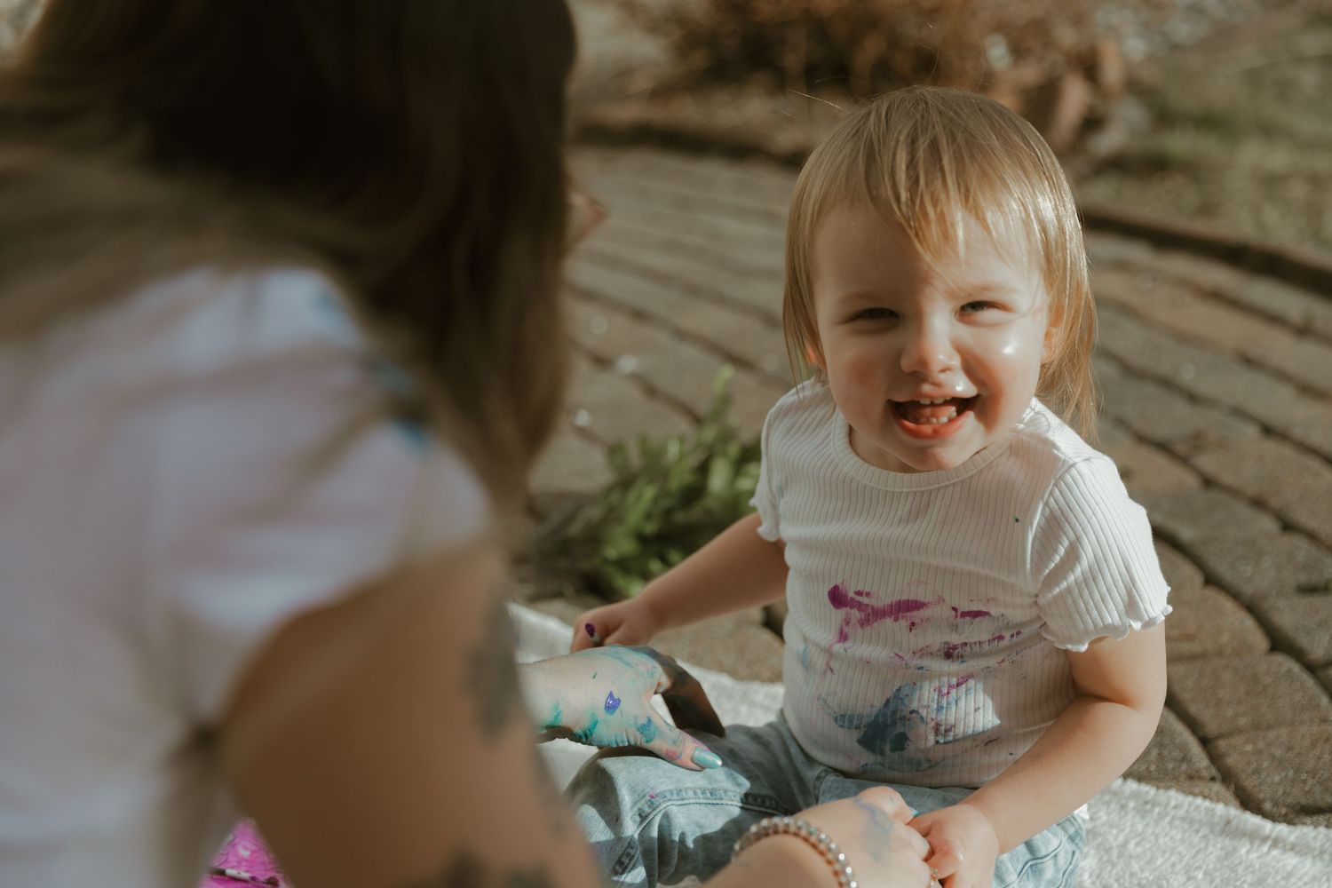 A small child in blue jeans and white t-shirt sits on a stone patio, laughing with joy.