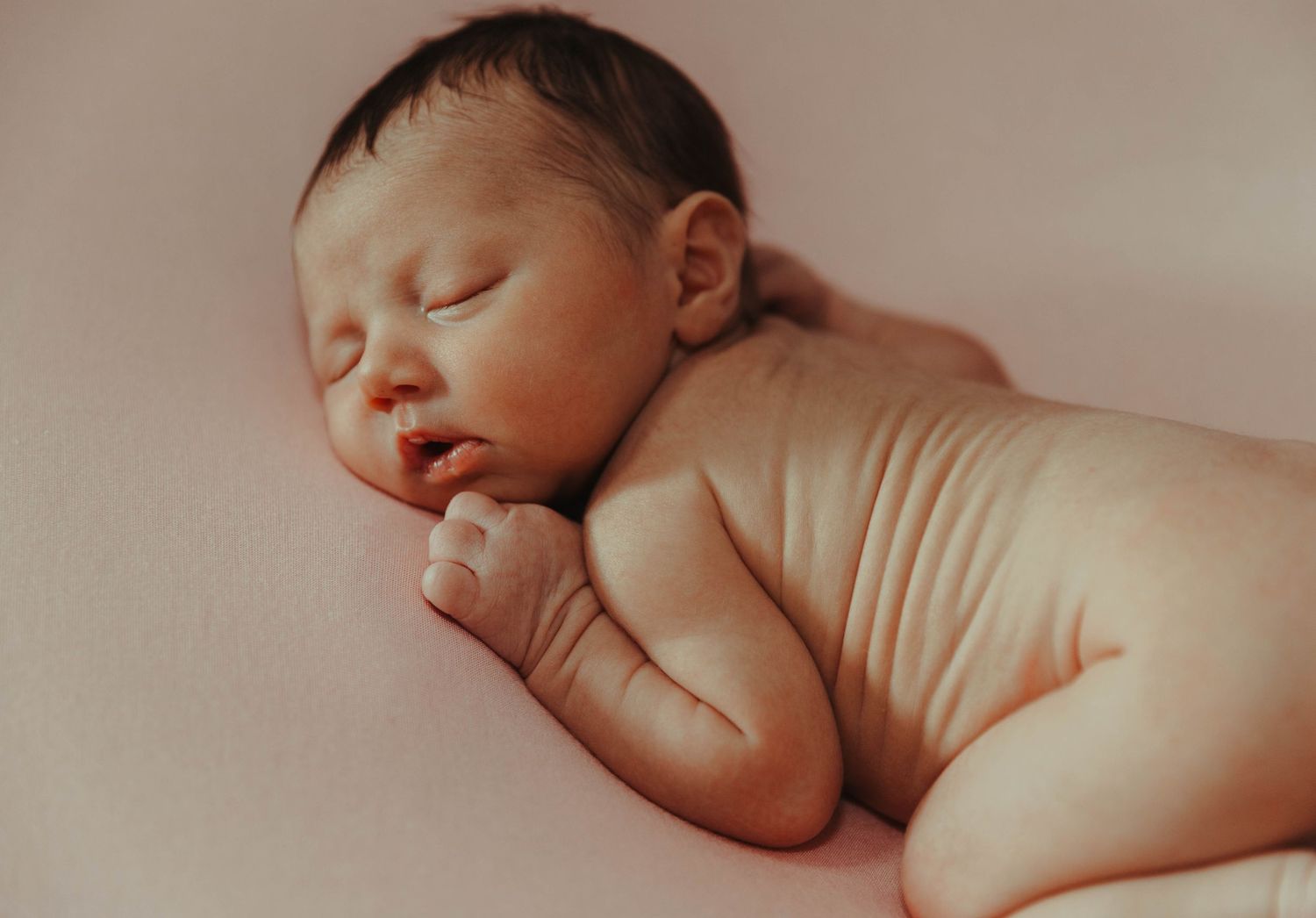 Peaceful sleeping newborn baby lying on soft pink fabric in natural light with curled fingers.